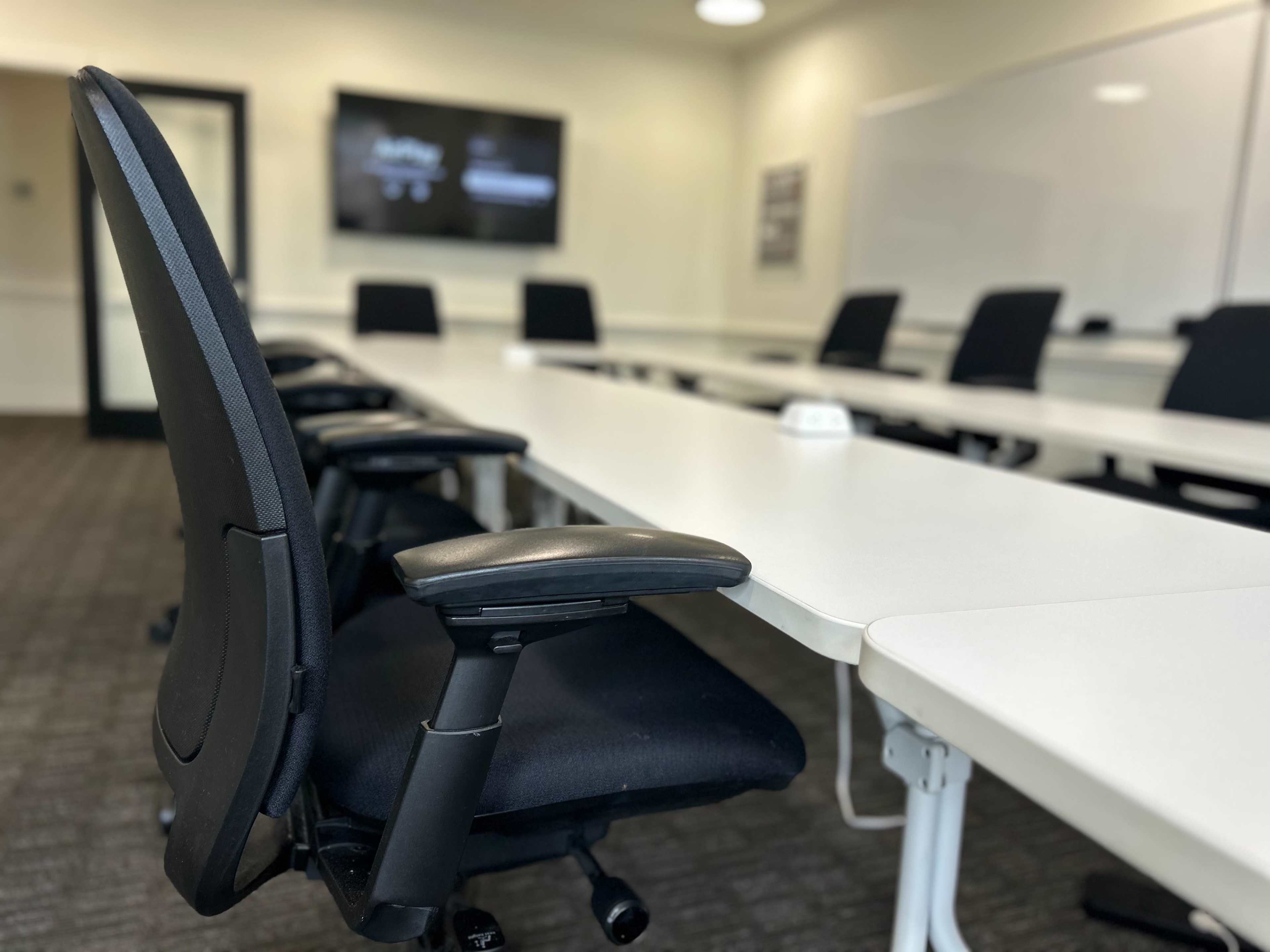 The image shows a modern conference room with a long white table, several black chairs, and a large screen mounted on the wall.