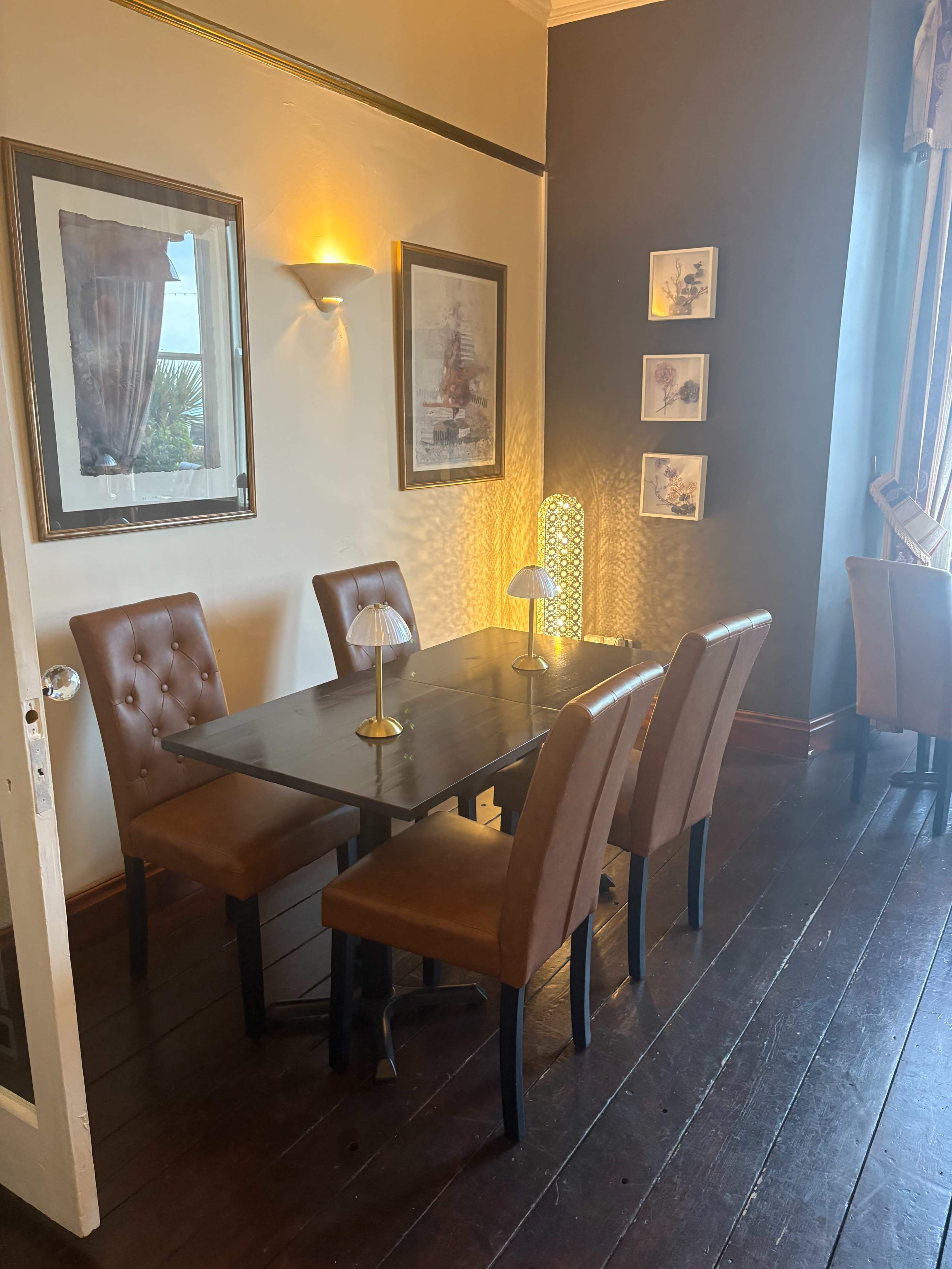 A small dining area features a wooden table with four brown upholstered chairs and is illuminated by wall sconces and a table lamp.