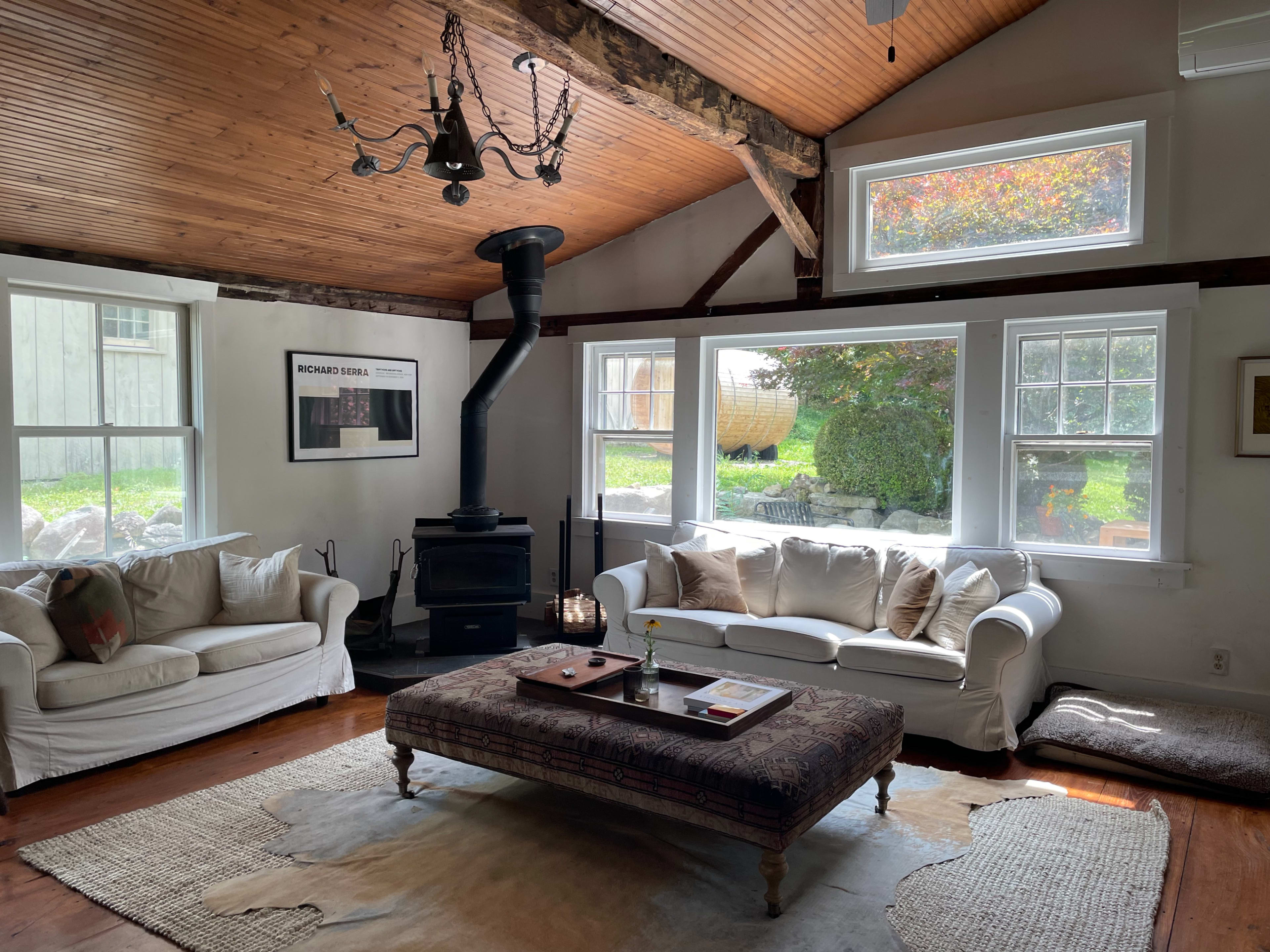 The living room features two white sofas, a central coffee table, and a wood stove, with large windows allowing natural light to illuminate the space.