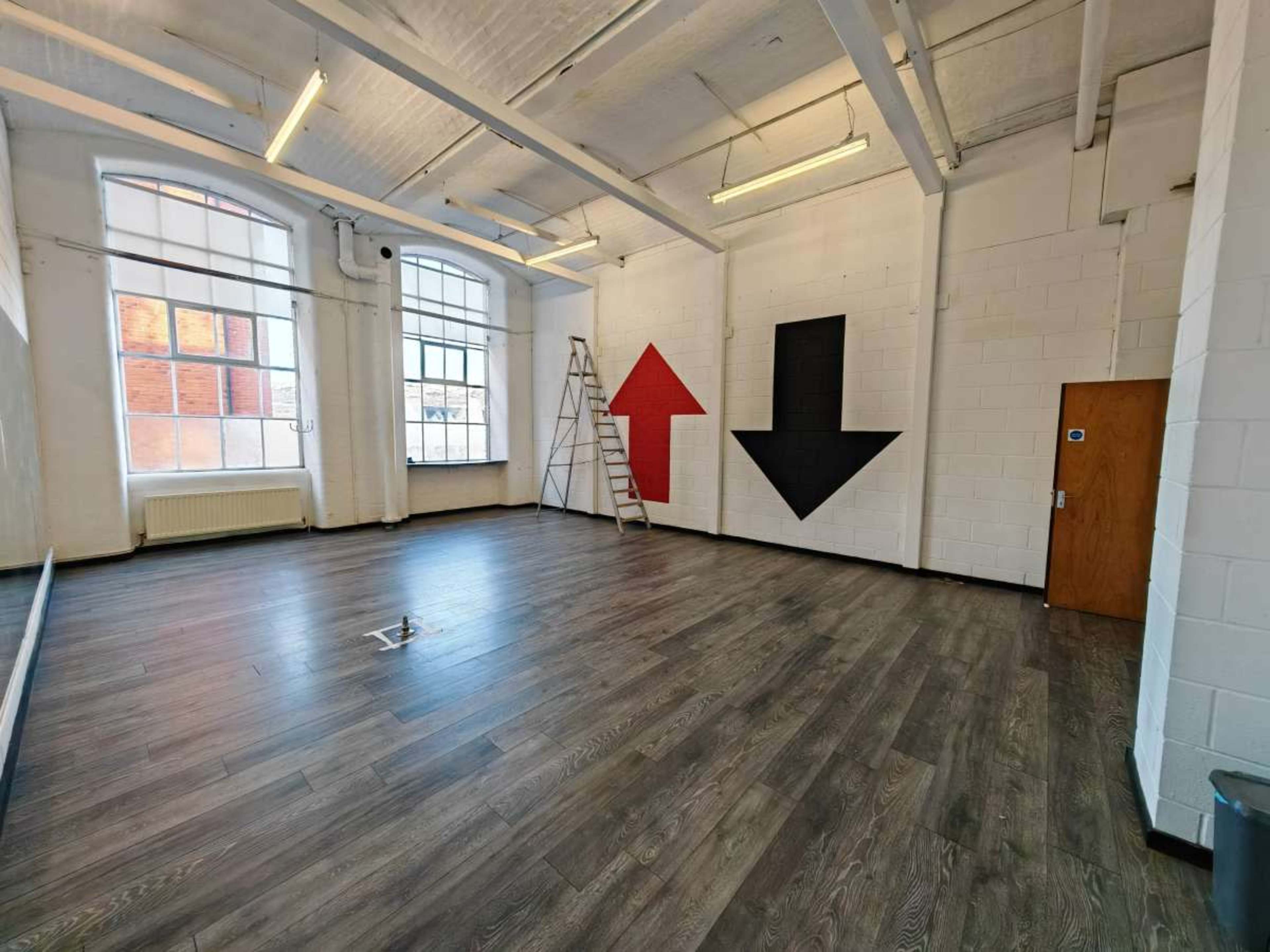 The image shows an empty room with wooden flooring, a ladder, and large red and black arrows painted on the white walls.