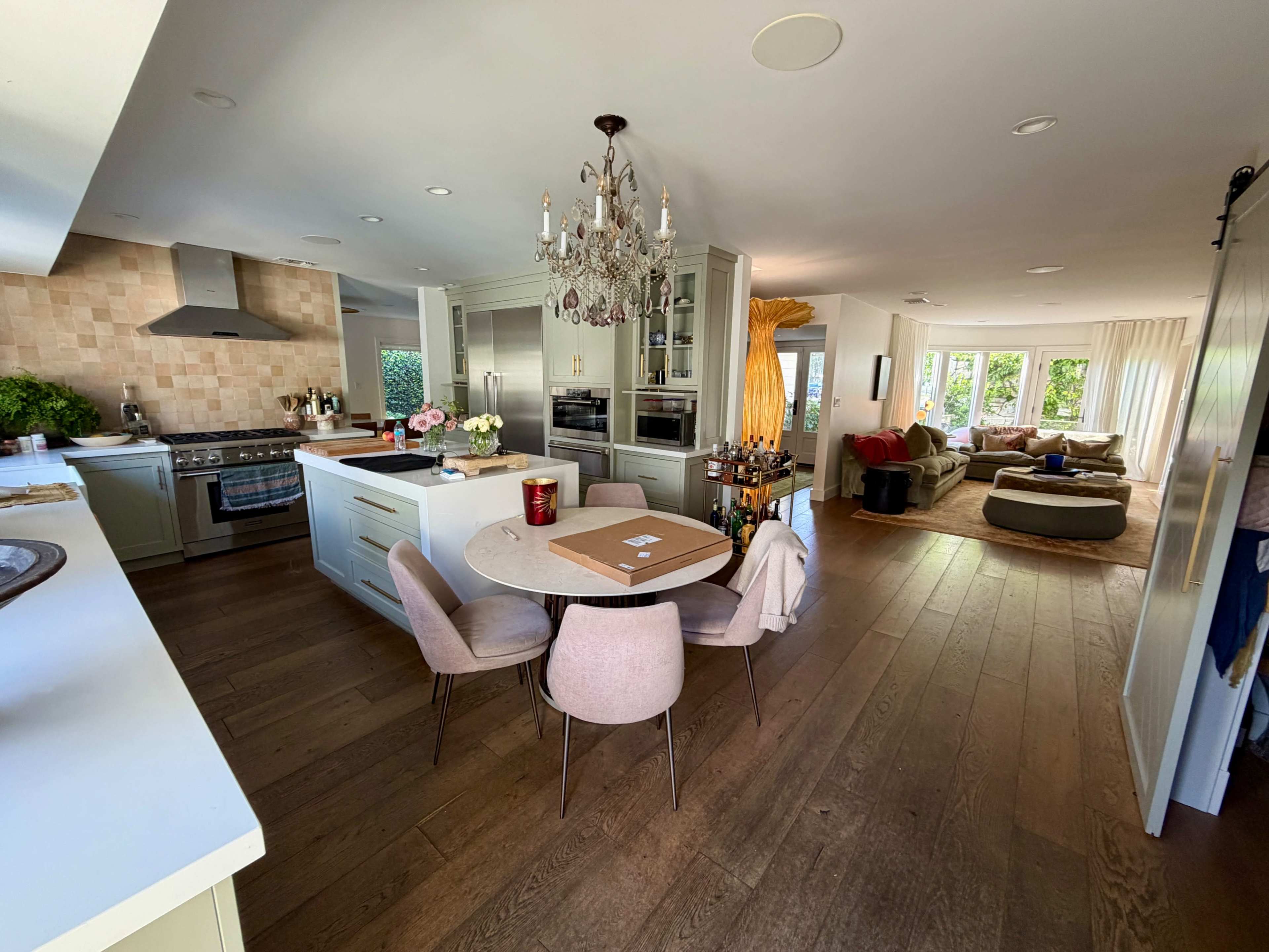 The image shows a modern kitchen and living area with a round dining table, a chandelier, and a sectional sofa in a spacious layout.