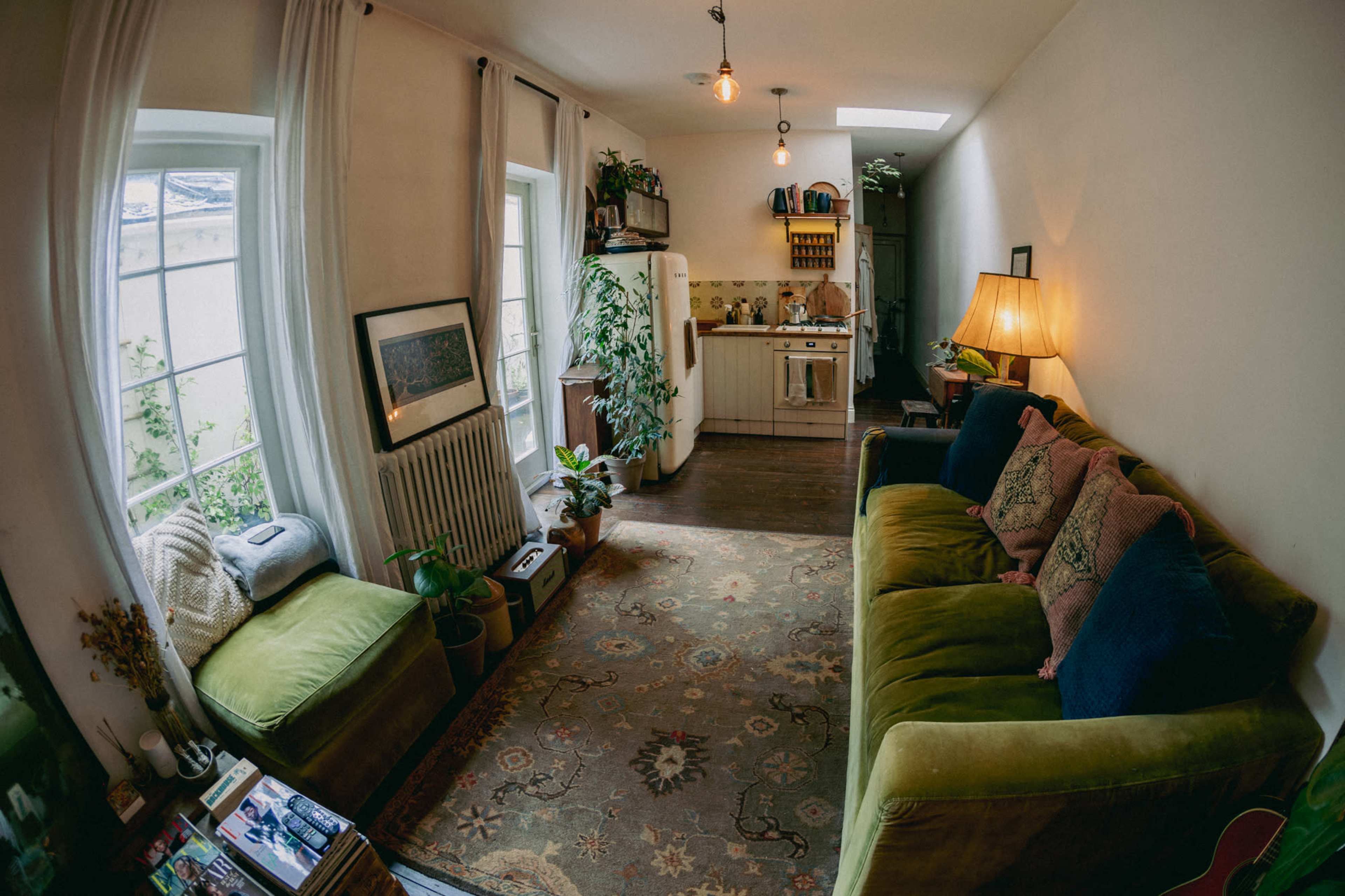 A cozy living room with a green sofa, a patterned rug, and a small kitchen area in the background, illuminated by natural light from the windows.