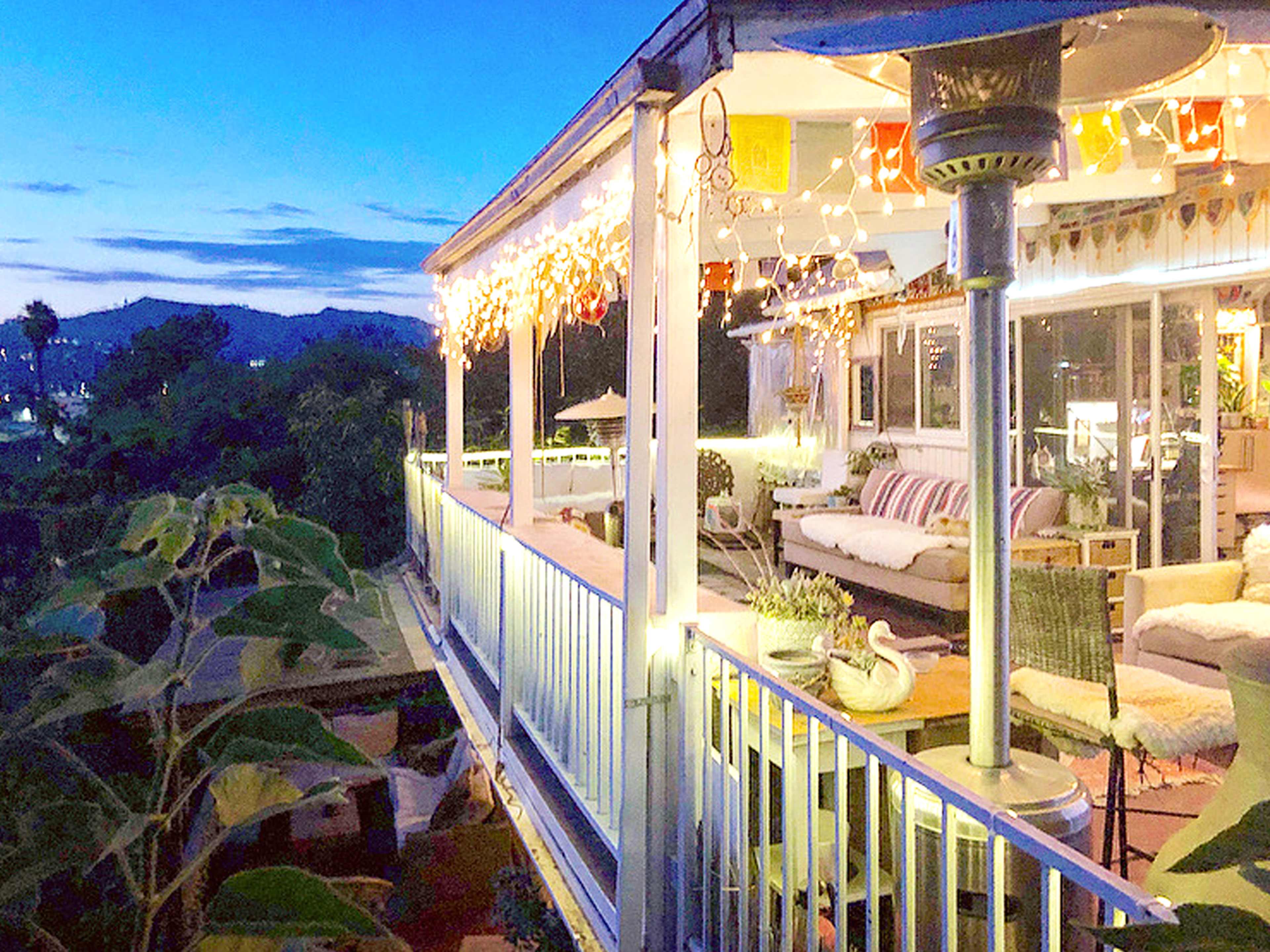 A patio is illuminated with string lights and decorated furniture, overlooking a scenic view at dusk.