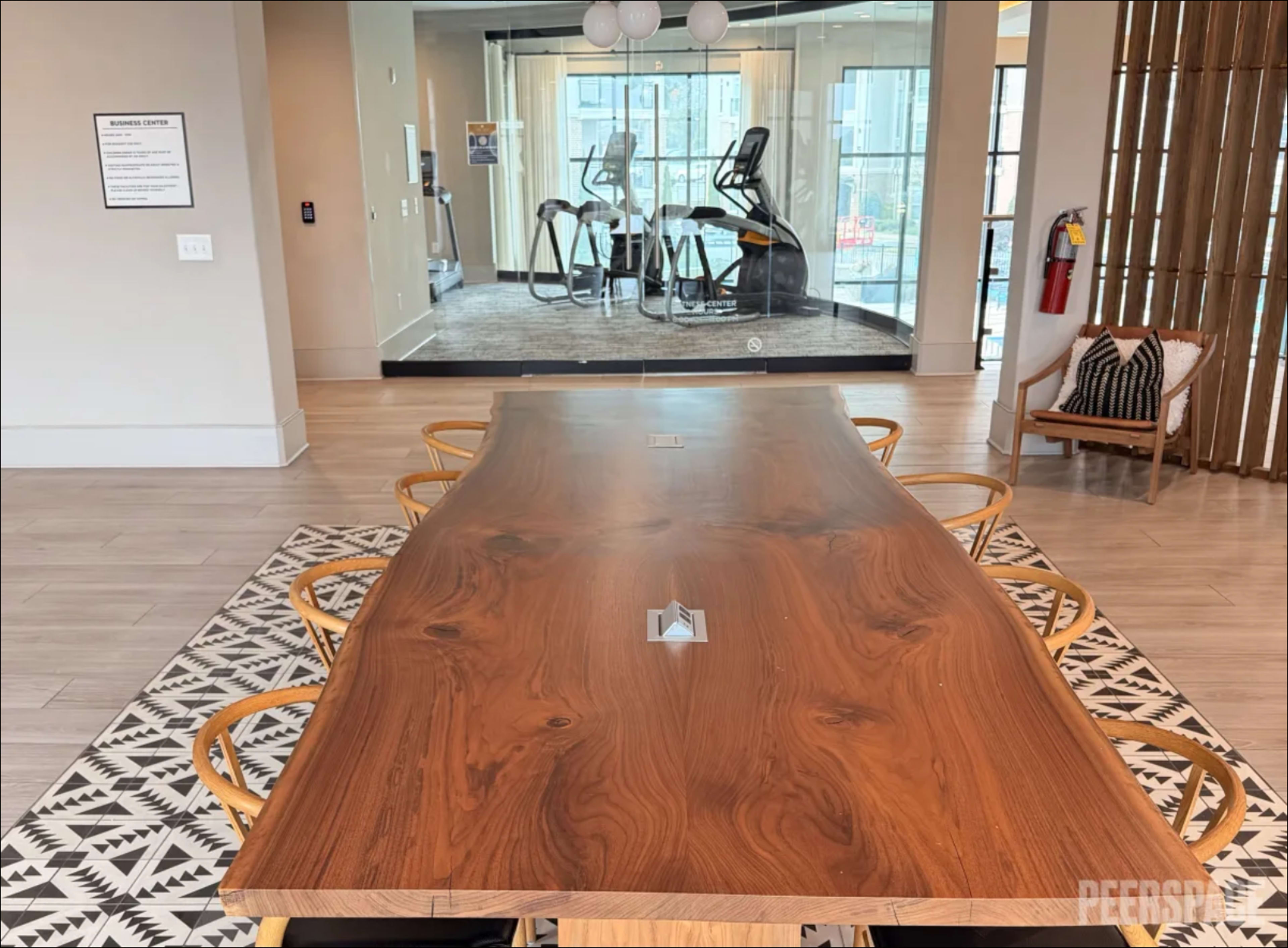A large wooden conference table with several chairs is in the foreground, while a fitness area with exercise equipment is visible through glass in the background.