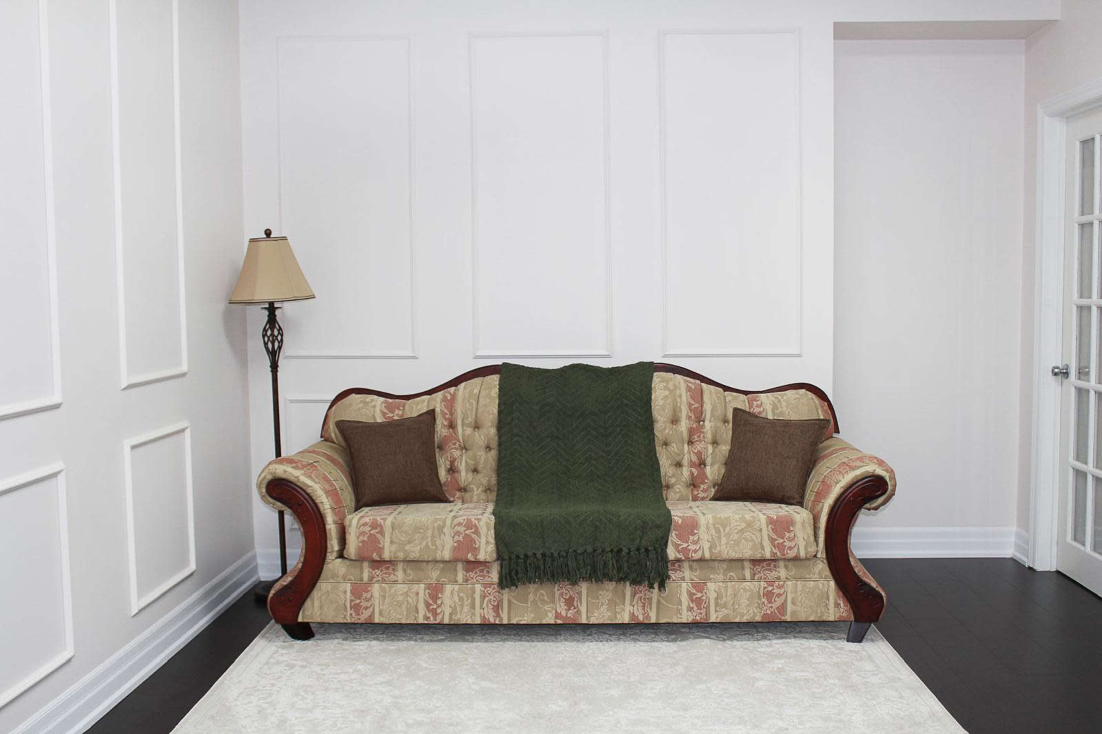 A floral-patterned couch with a green throw blanket and two brown cushions sits in a minimalist room with white walls and a lamp in the corner.