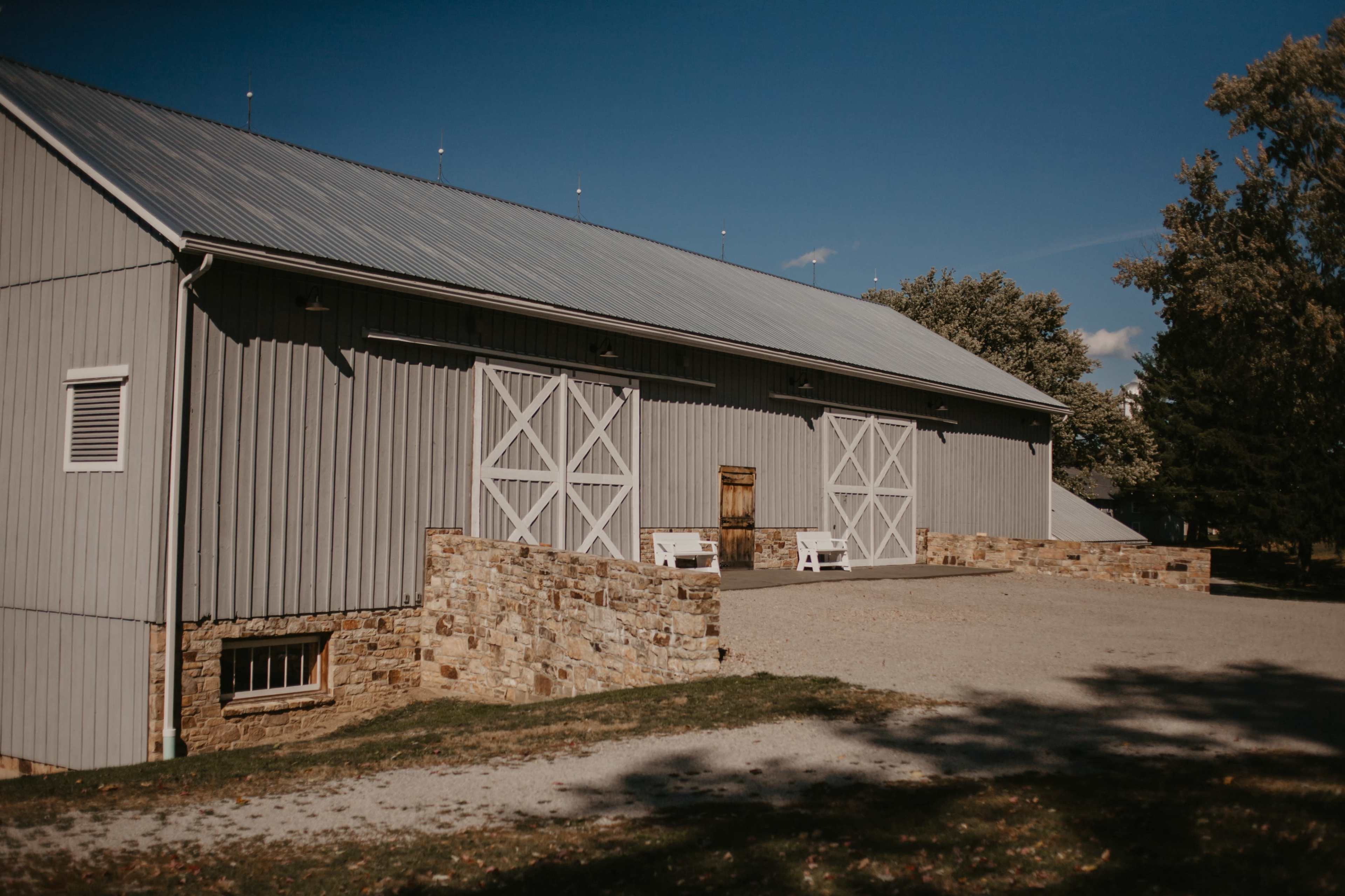 A gray barn with white double doors stands on a gravel driveway surrounded by stone walls and trees under a clear blue sky.
