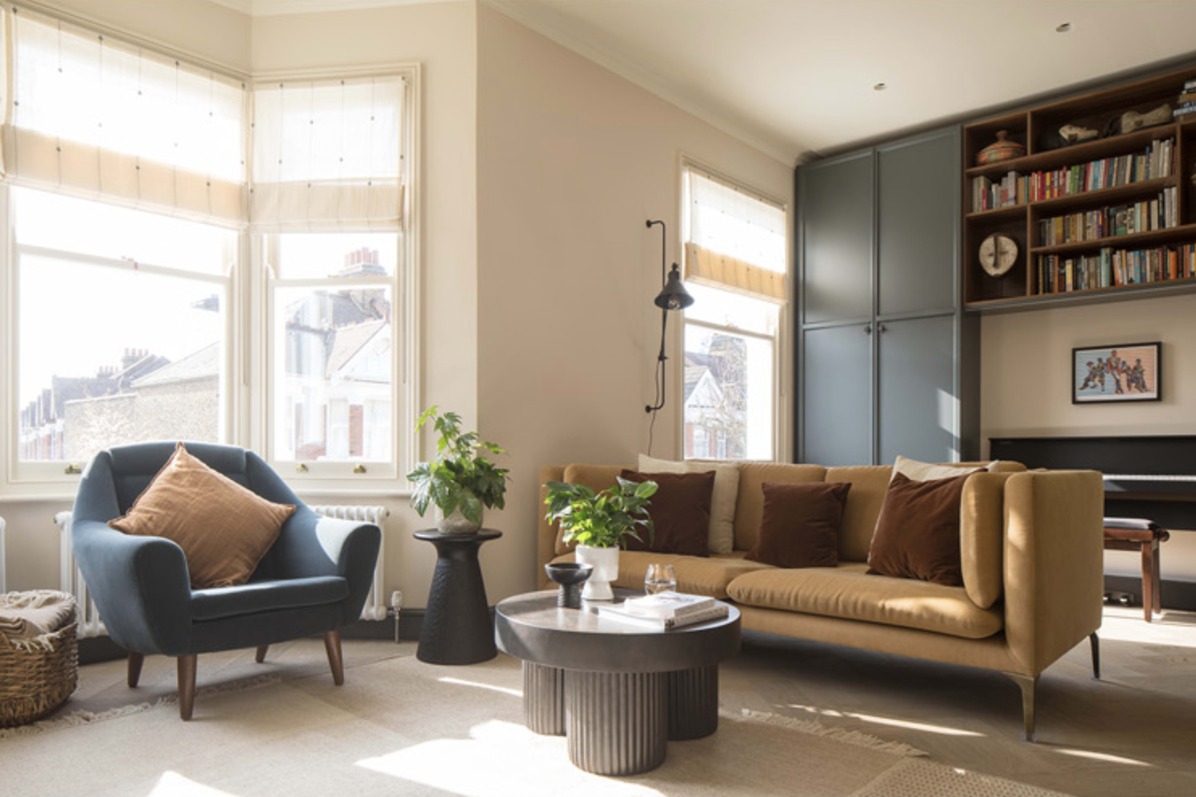 A cozy living room with a blue armchair, a tan sofa with decorative pillows, a circular coffee table, and bookshelves in the background.