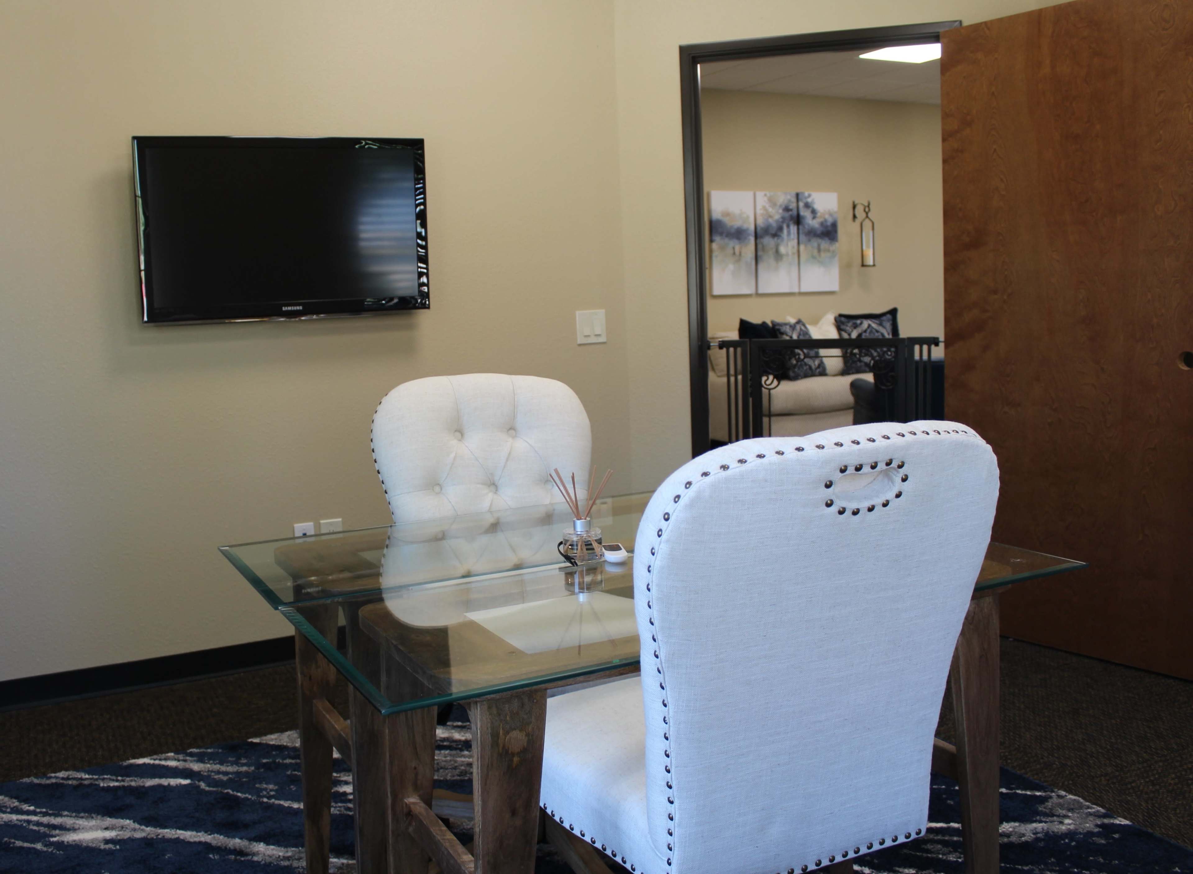 A glass-top table with two upholstered chairs is positioned in a small office space featuring a wall-mounted television and a doorway leading to another room.