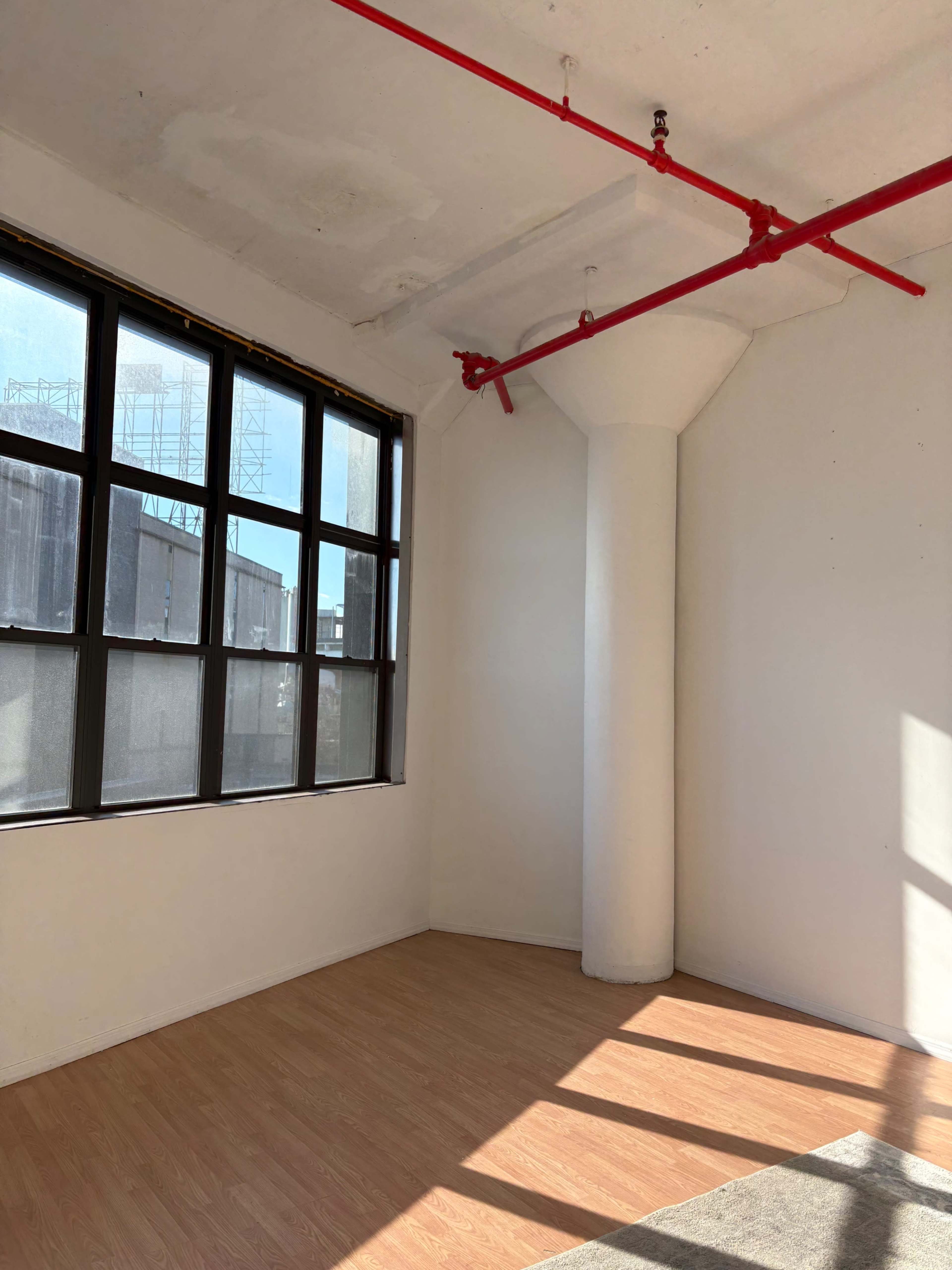 The image shows an empty room with a large window, a red pipe running along the ceiling, and light-colored wooden flooring.