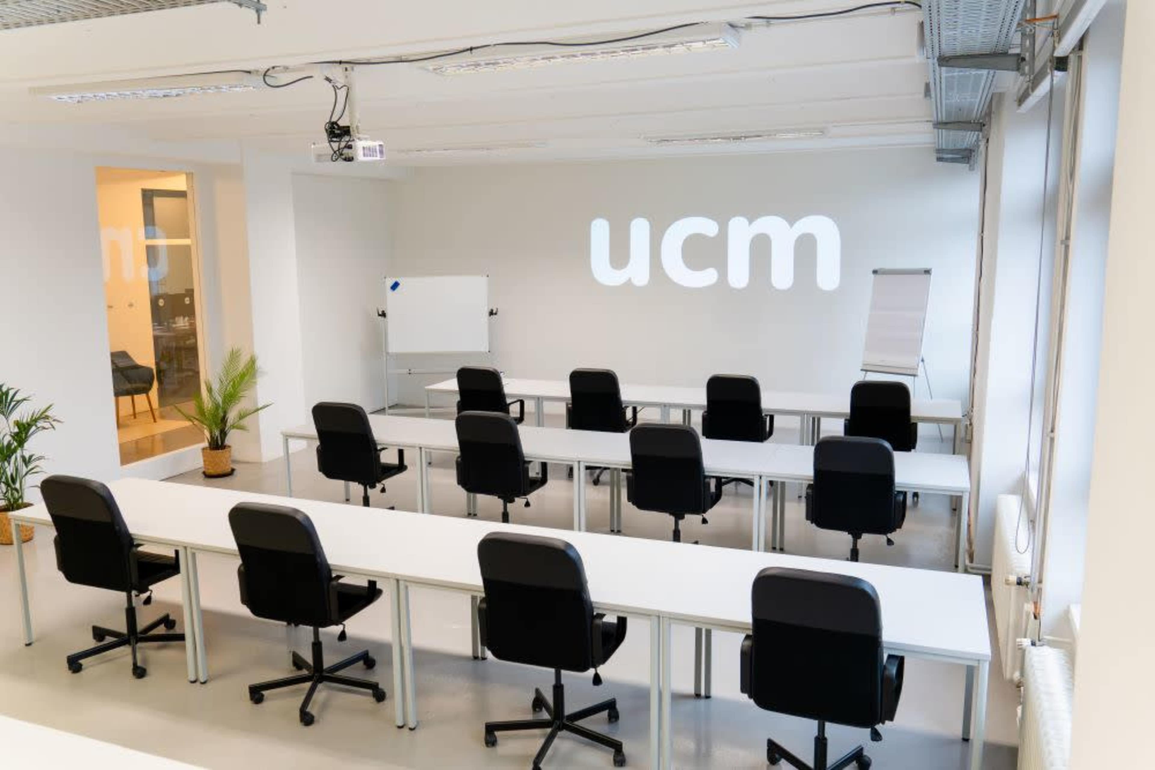 The image shows a modern classroom with multiple rows of black chairs and white tables arranged facing a wall displaying the text "ucm."