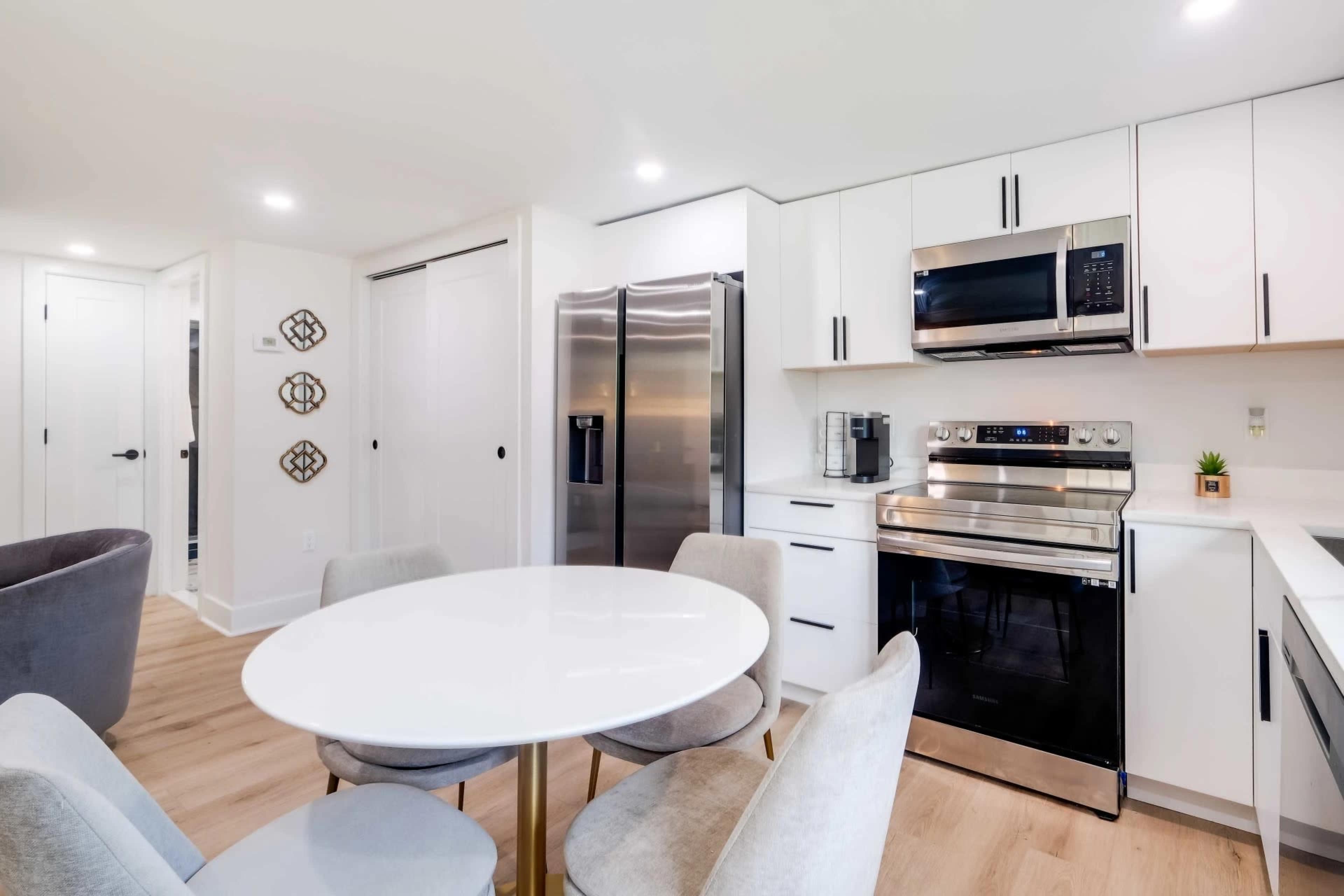 A modern kitchen features a round white table surrounded by gray chairs, stainless steel appliances, and sleek, white cabinetry.