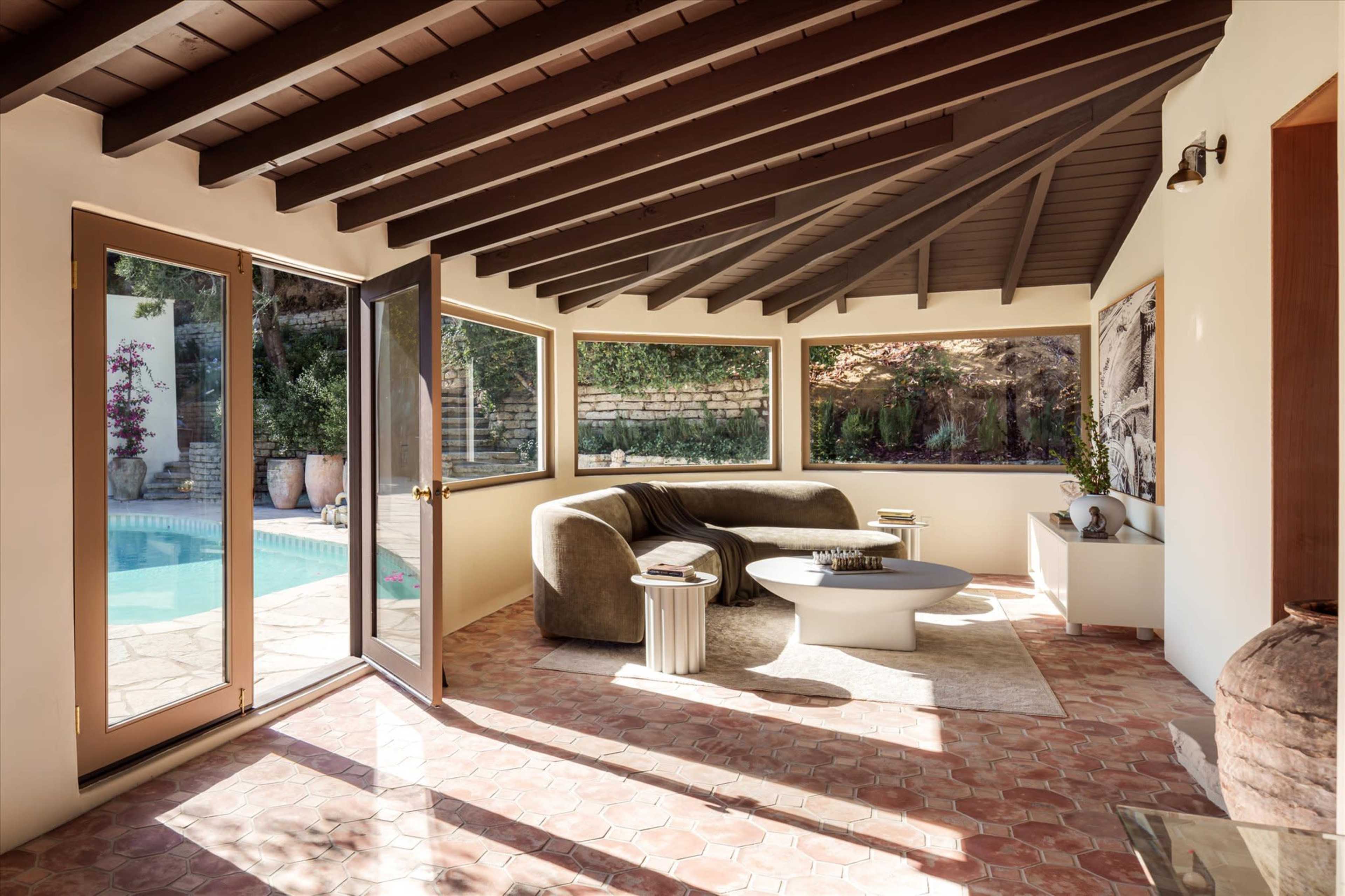 A modern living room features large windows overlooking a pool, with a curved couch and minimalist furnishings.