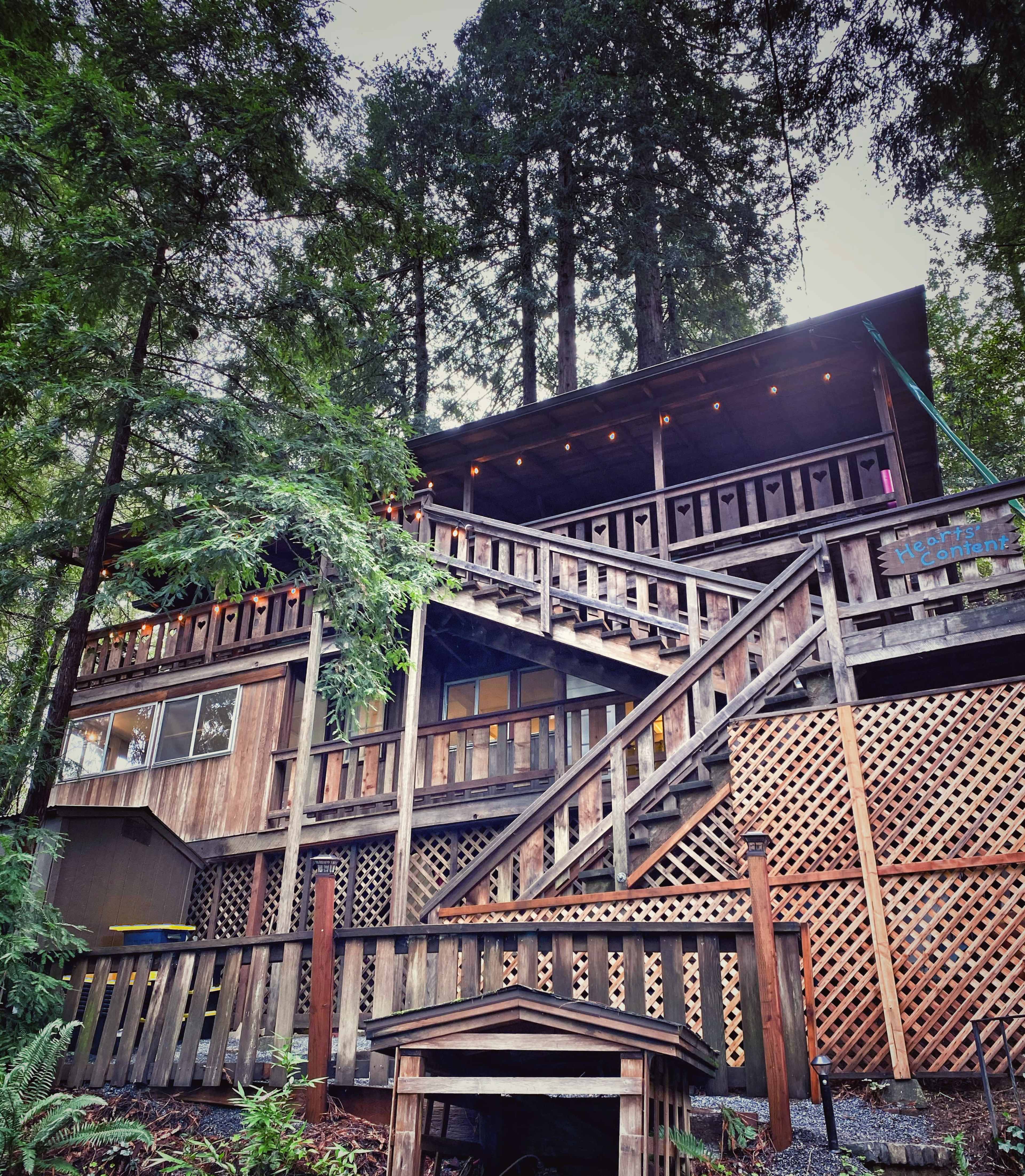 A multi-level wooden cabin built among tall trees features multiple terraces and an intricate staircase leading to the main entrance.