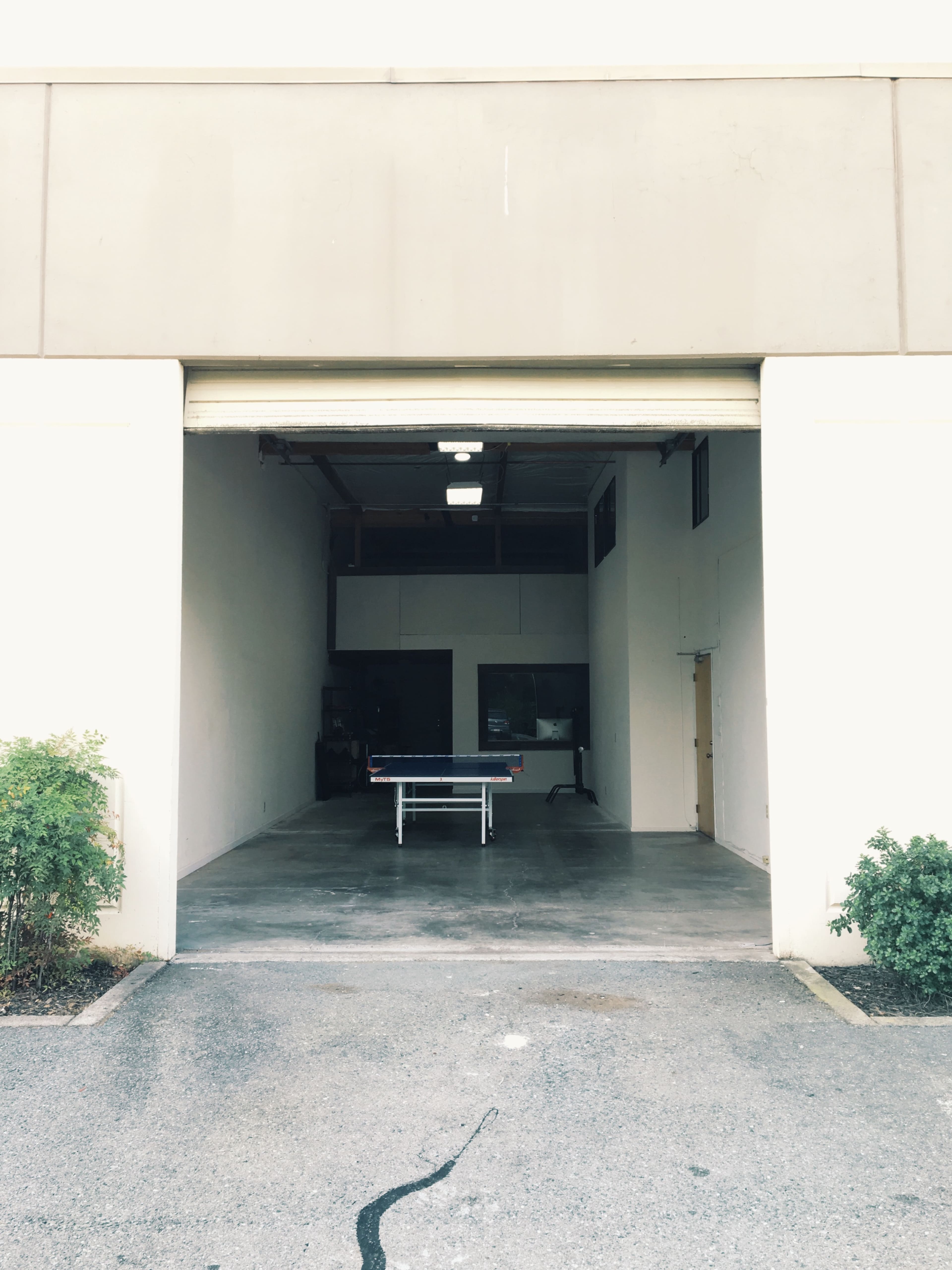 A large open garage with a ping pong table positioned inside, flanked by low shrubs outside.
