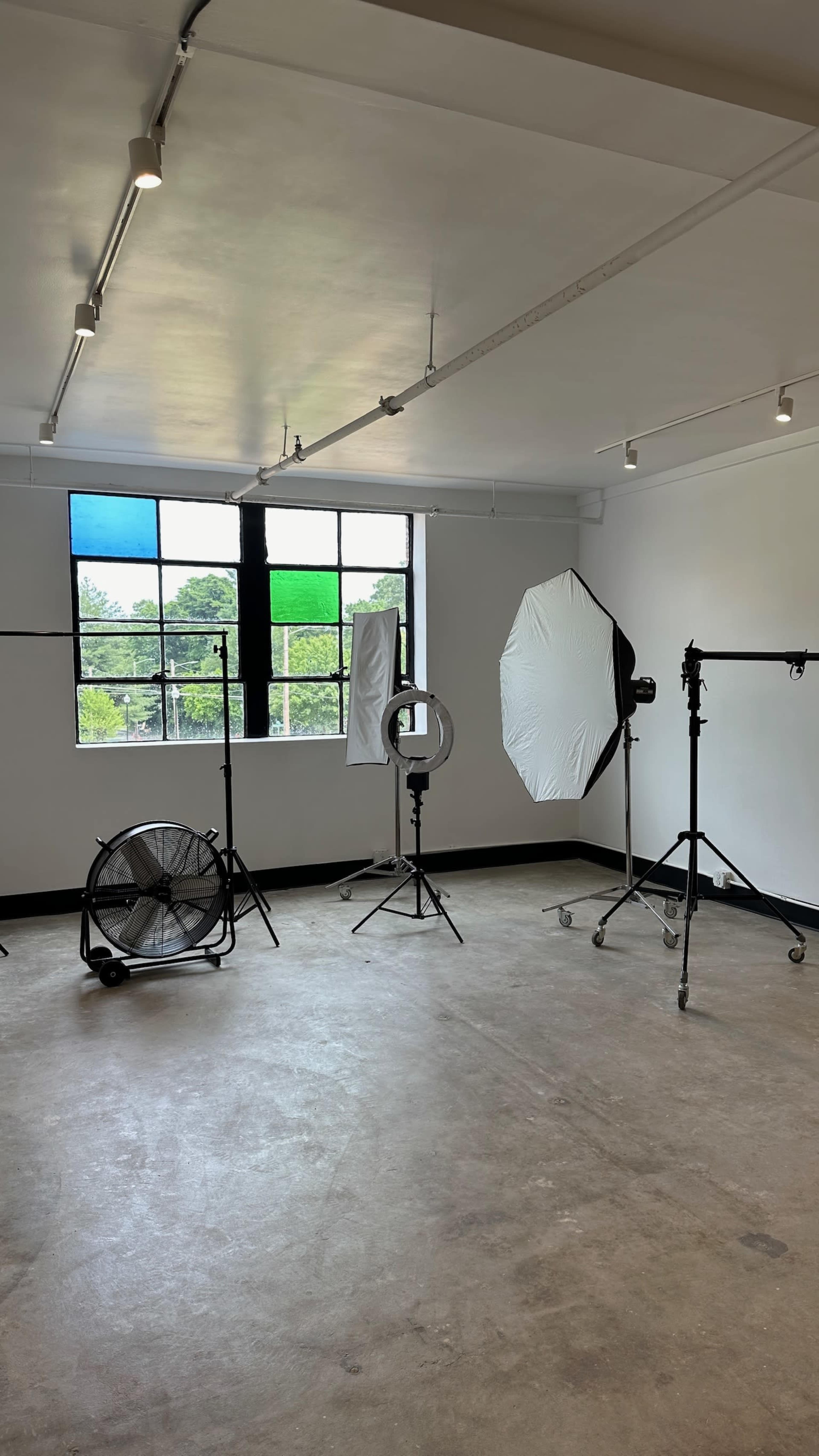 A photography studio with lighting equipment, backdrops, and a fan positioned near large windows.