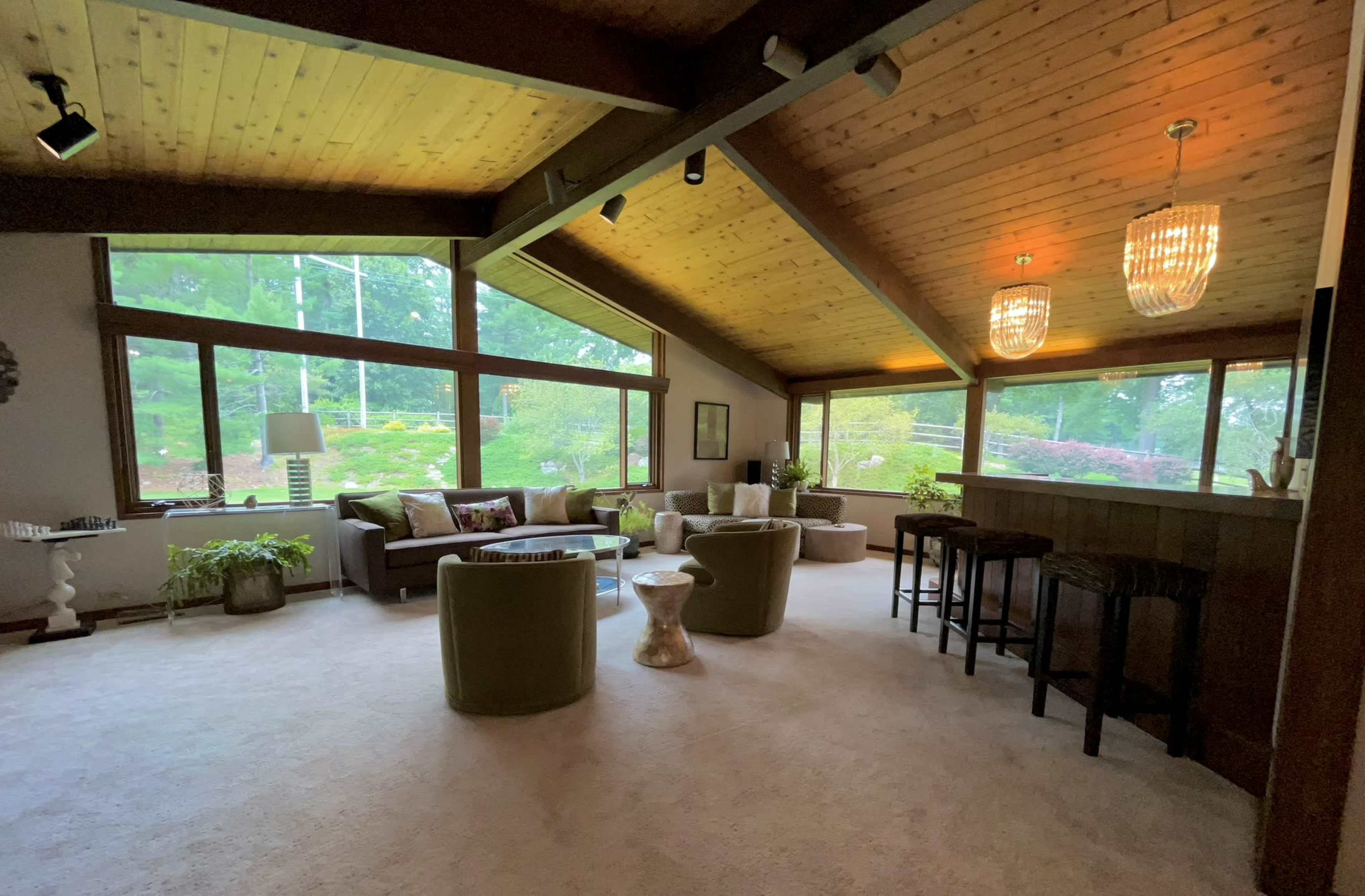 The image shows a spacious living room with large windows, wooden beams, and a bar area featuring stools.