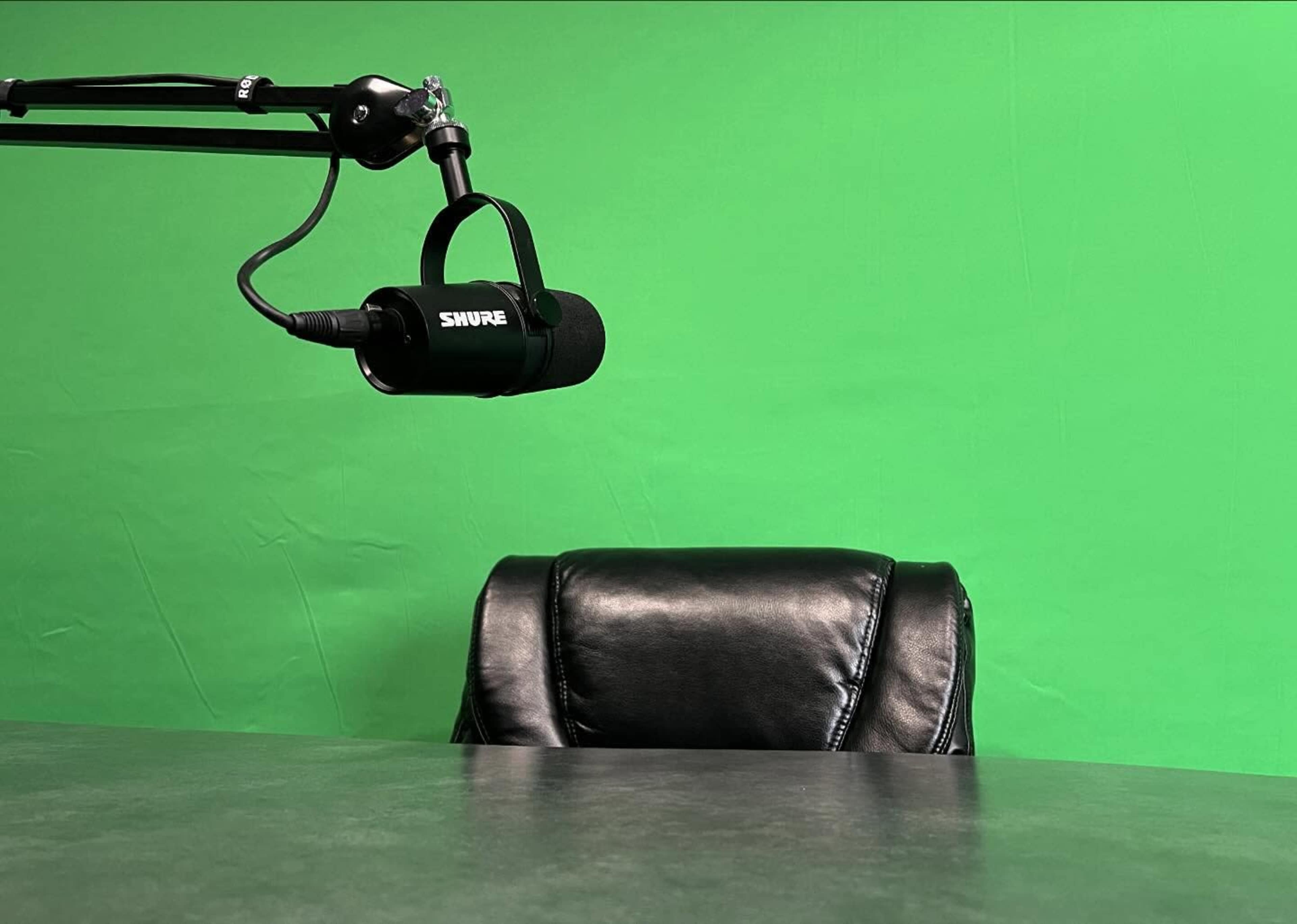 A black leather chair sits empty at a table with a microphone mounted on an adjustable arm, all set against a green backdrop.