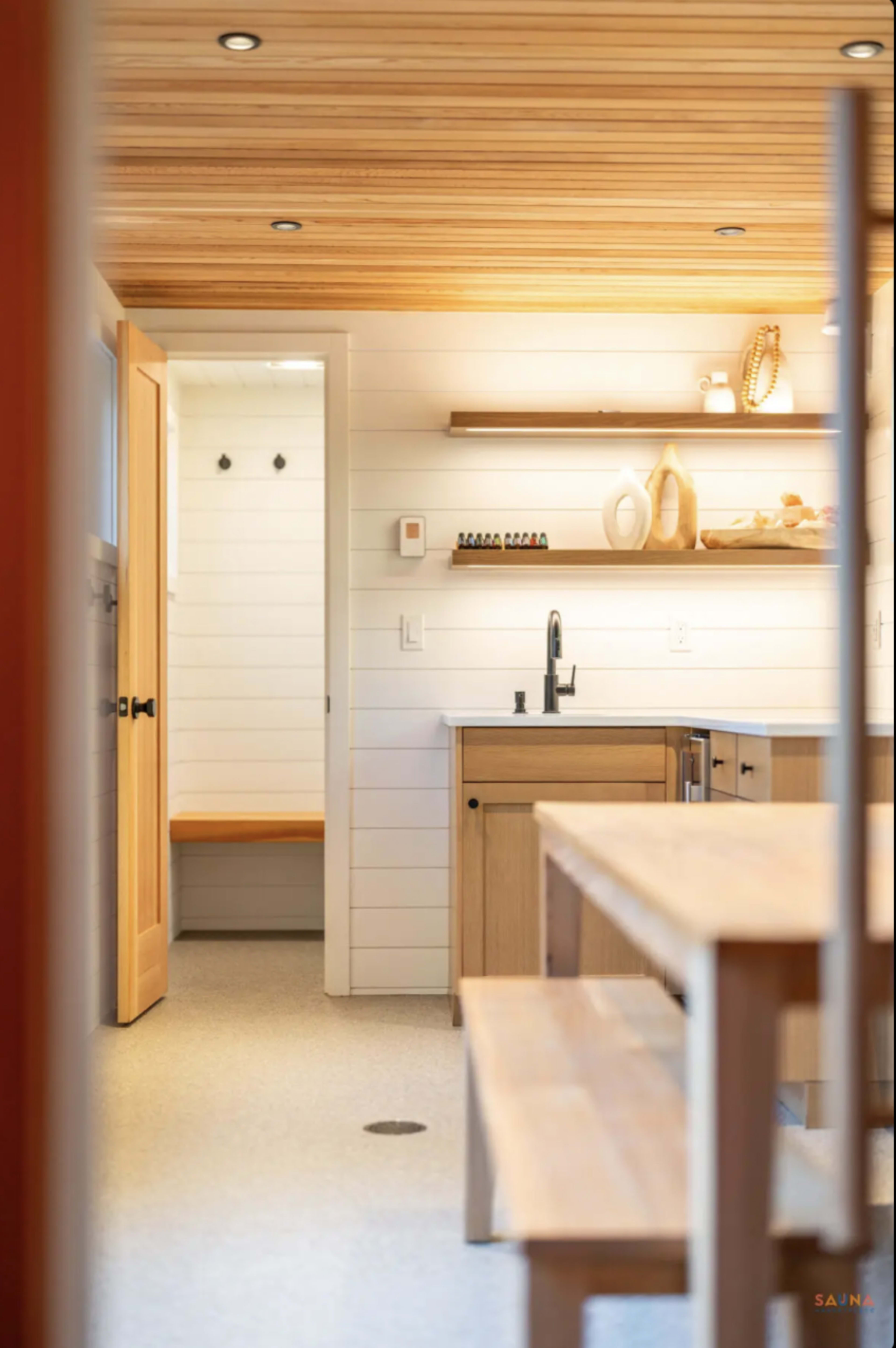 The image shows a modern kitchen with wooden countertops, white shiplap walls, and a view of a hallway leading to a room with hooks on the wall.