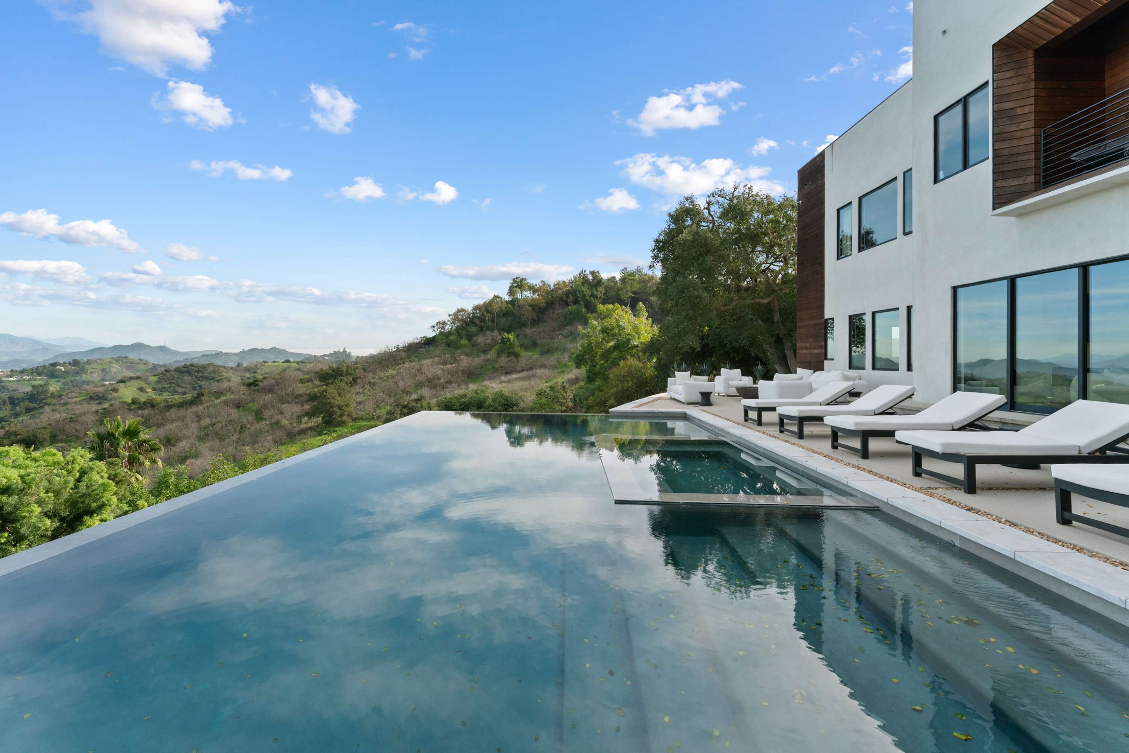 A modern villa features a sleek infinity pool overlooking a hilly landscape under a partly cloudy sky.