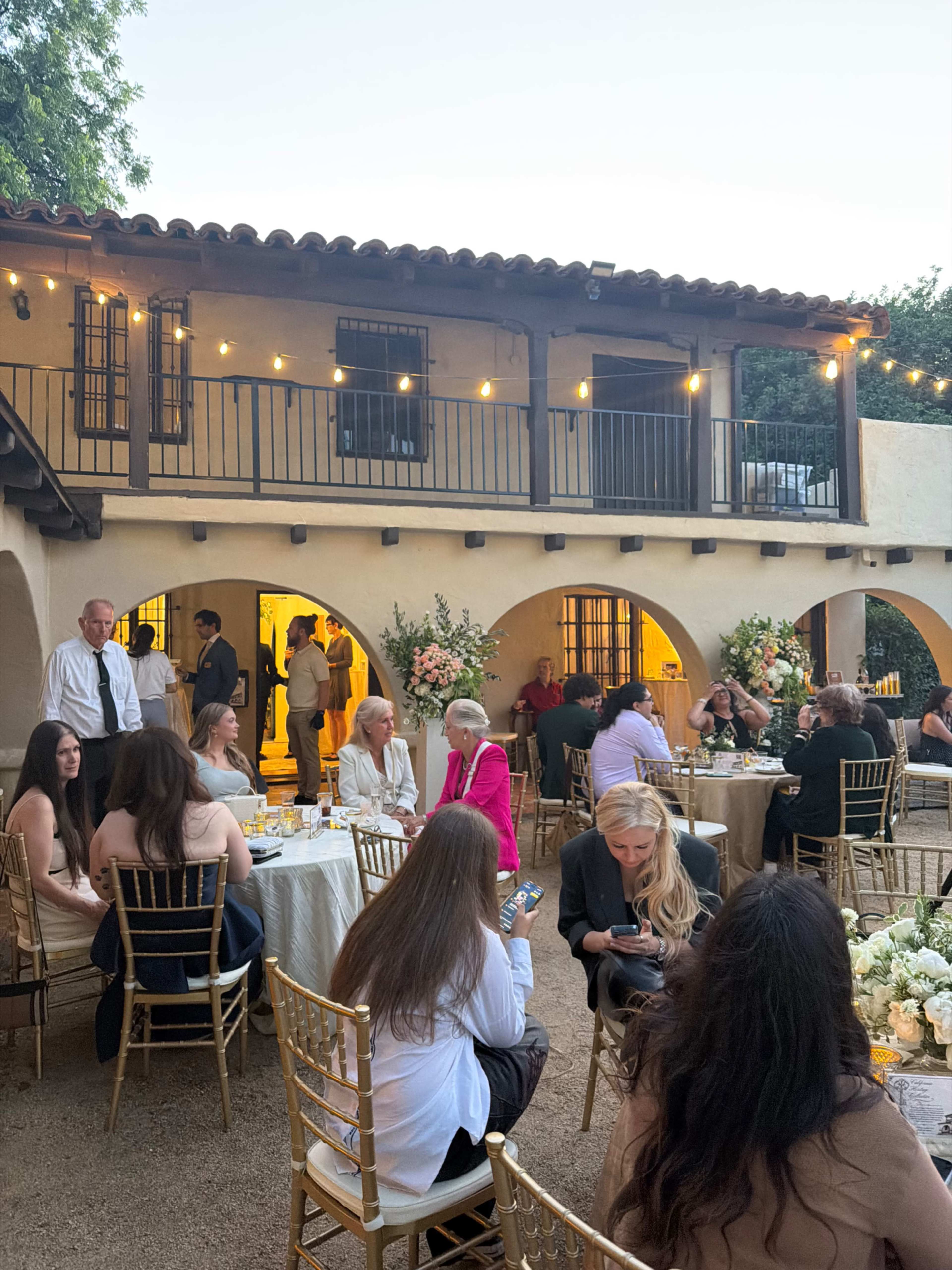 A group of people is gathered outside at an event, seated at tables with flowers and enjoying a warm evening ambiance under string lights near a building with a balcony.