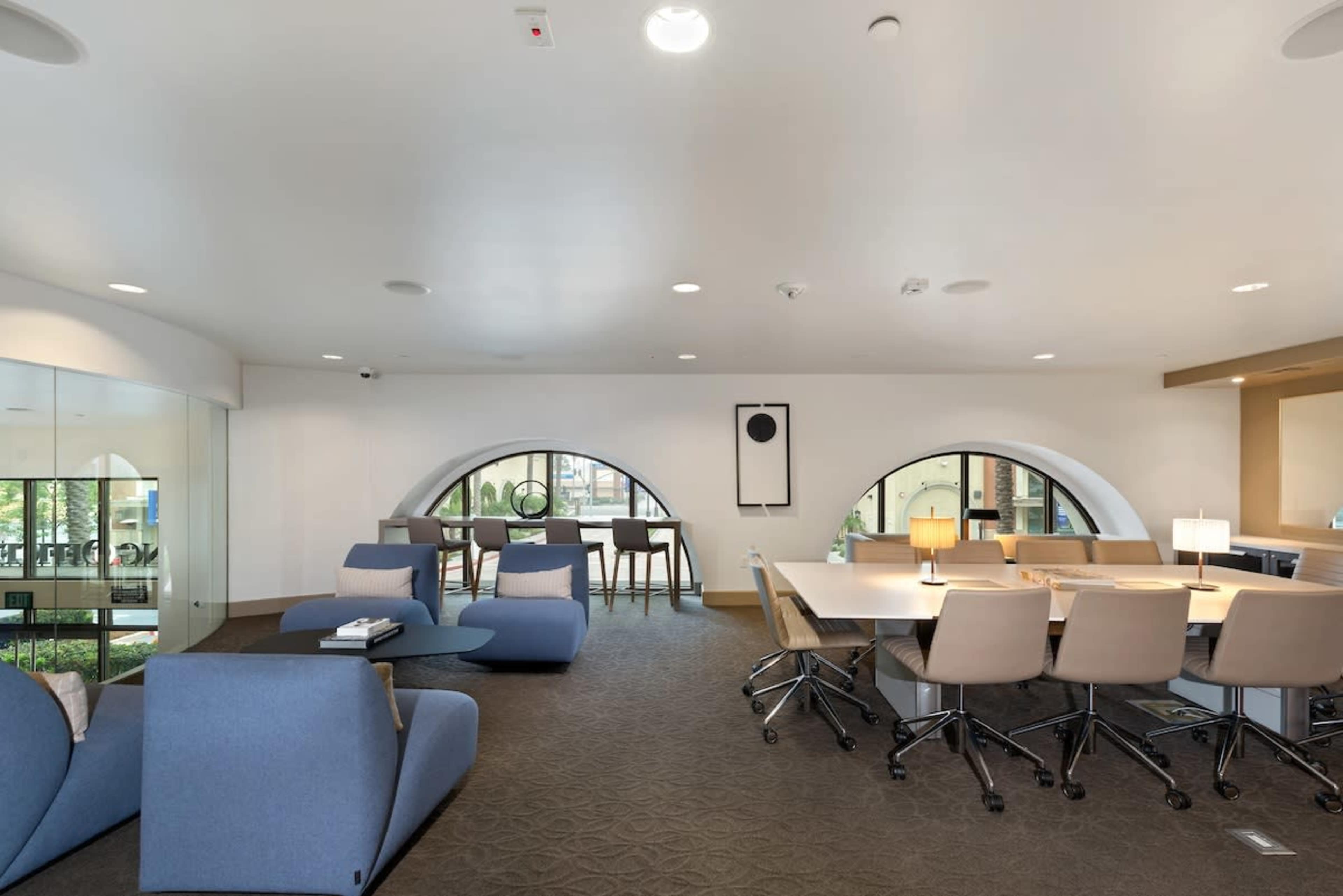 An office space with curved windows, a seating area with blue chairs, and a long table surrounded by light-colored chairs.