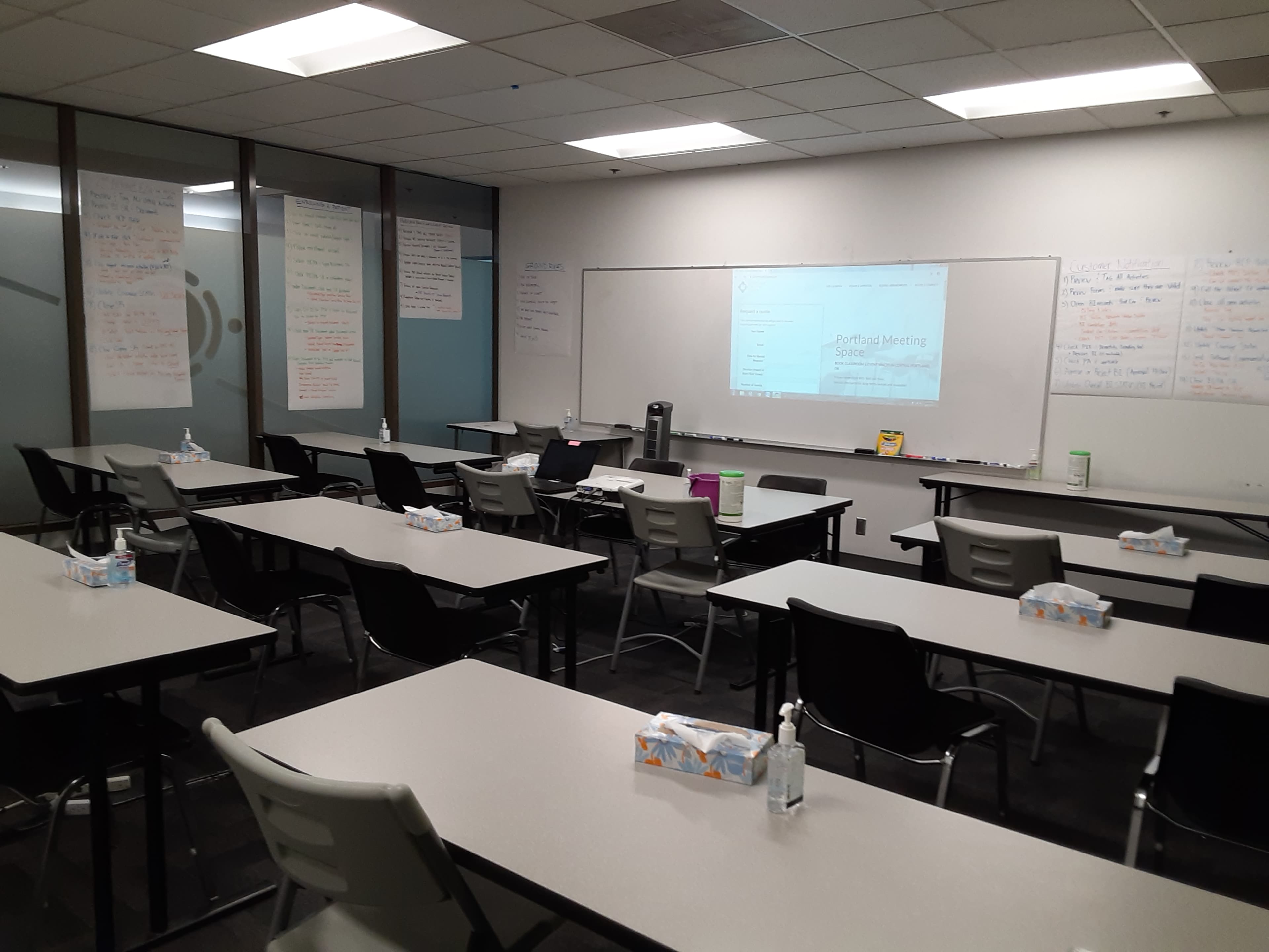 A classroom is set up with multiple tables and chairs arranged for a meeting, featuring flip charts and a whiteboard in the background.