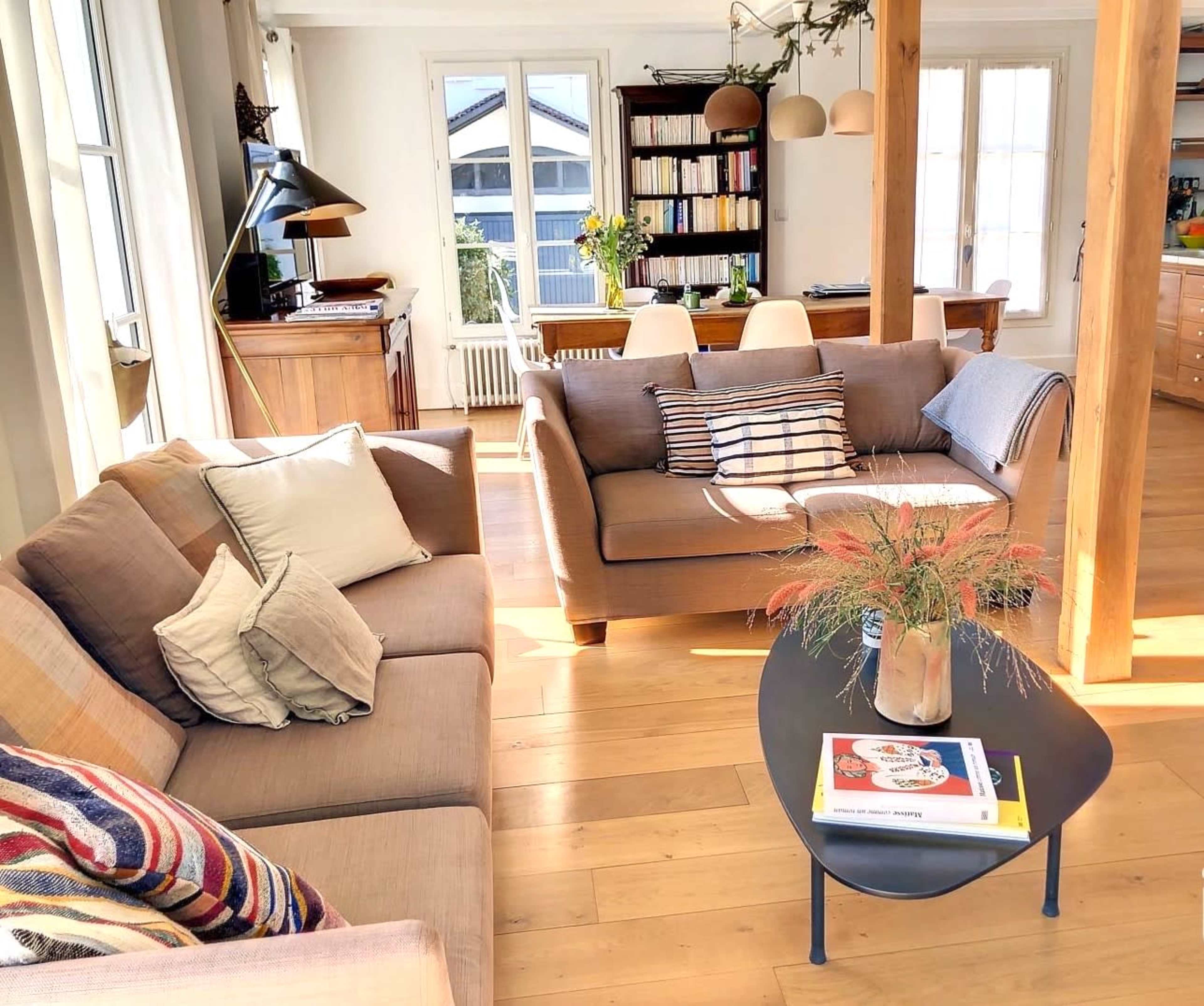 A bright living room with two sofas, a coffee table, and a dining area visible in the background, featuring wooden furniture and large windows.