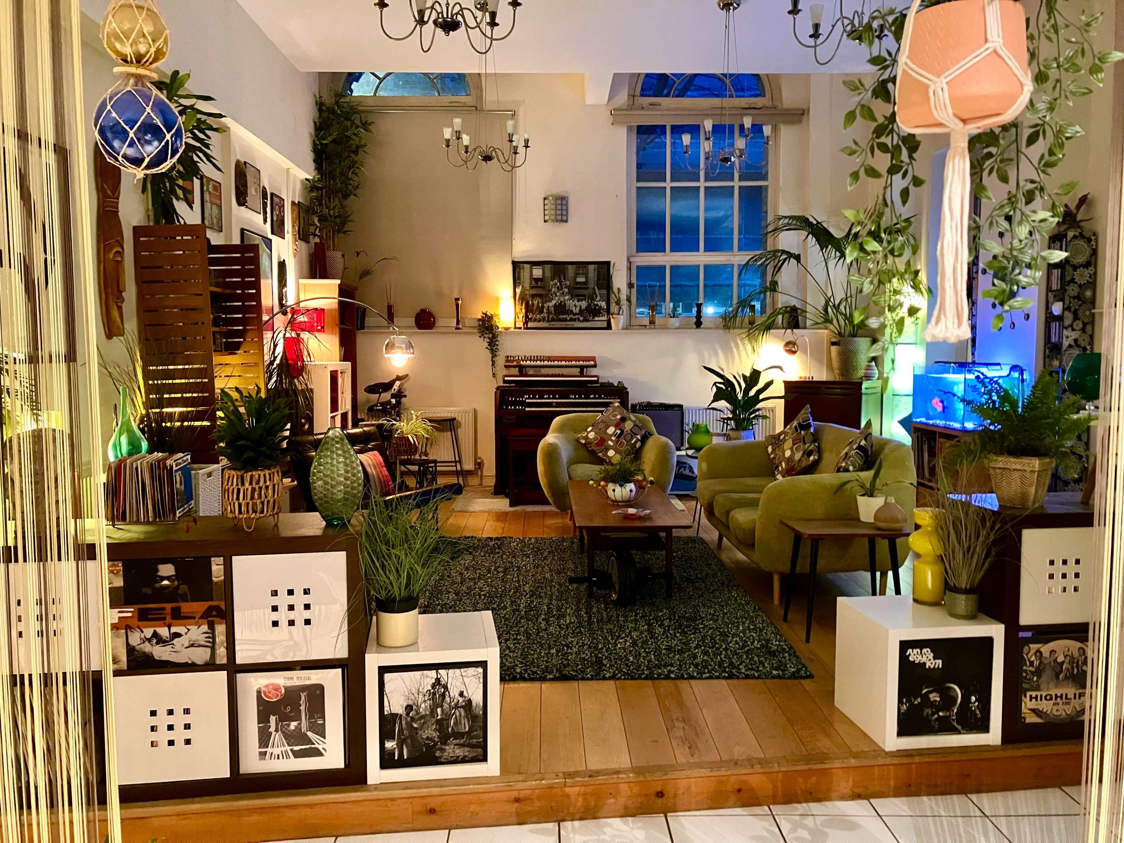 The scene features a cozy living room with green sofas, plants, and lit decorations, creating a welcoming atmosphere.