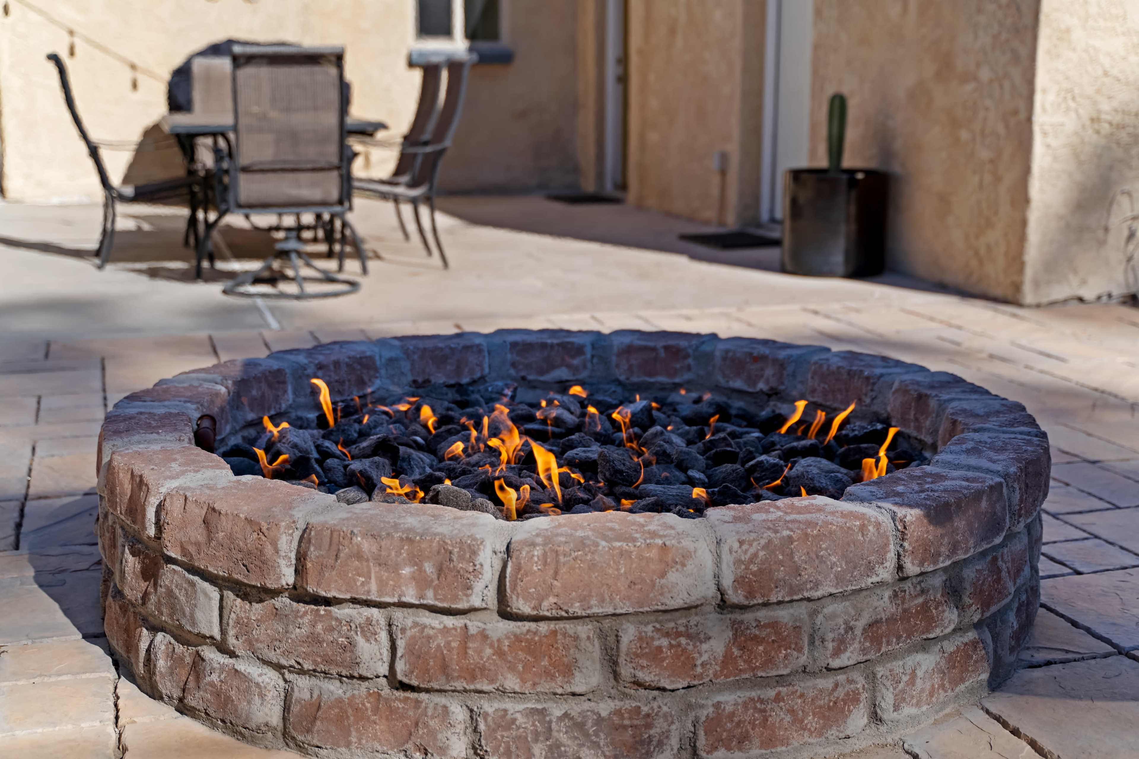 A circular stone fire pit with flames rising from charcoal sits on a patio next to outdoor seating.