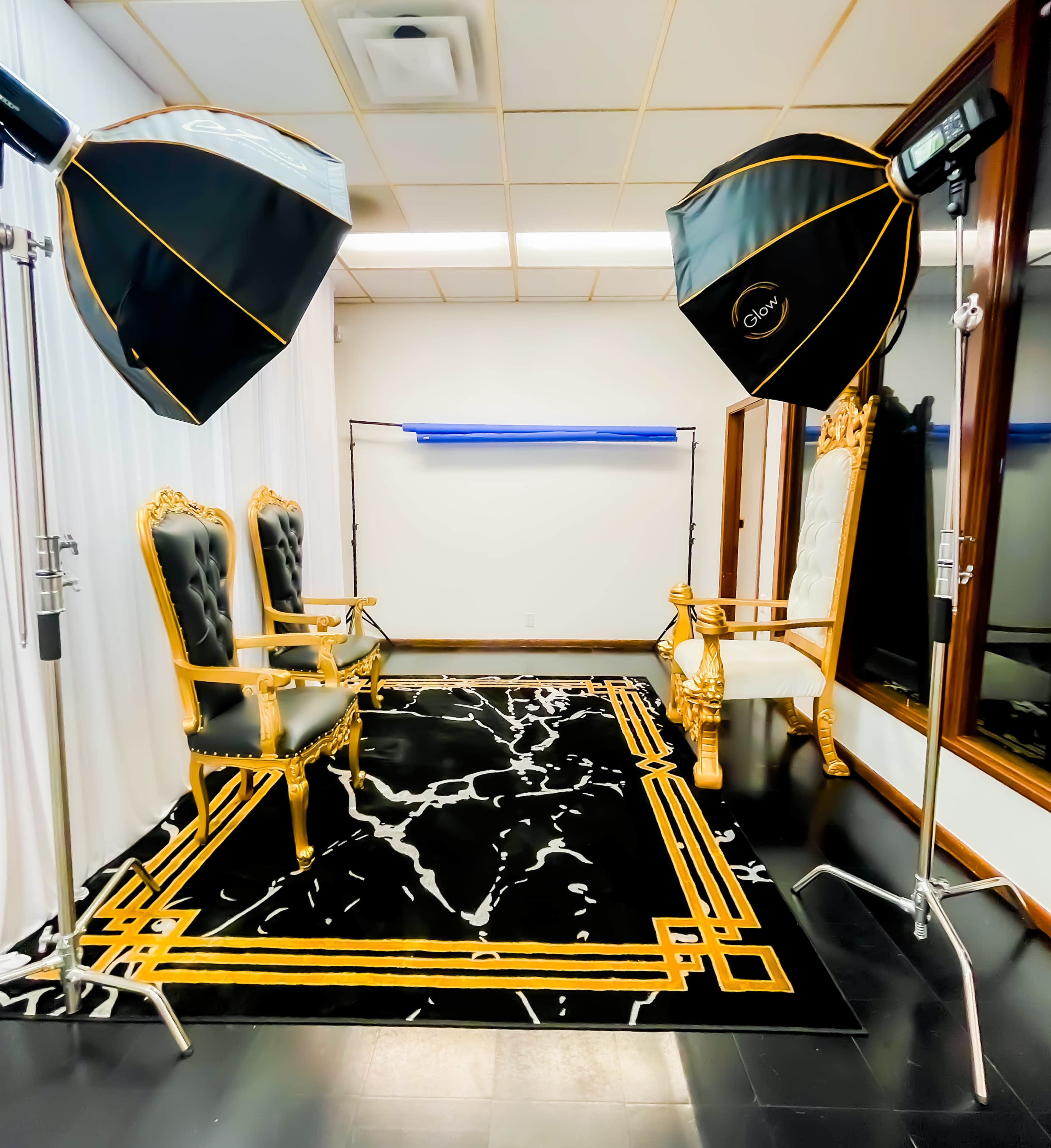 The image shows a photography studio setup with two golden chairs, a black and gold patterned rug, and two softbox lighting devices positioned on either side.