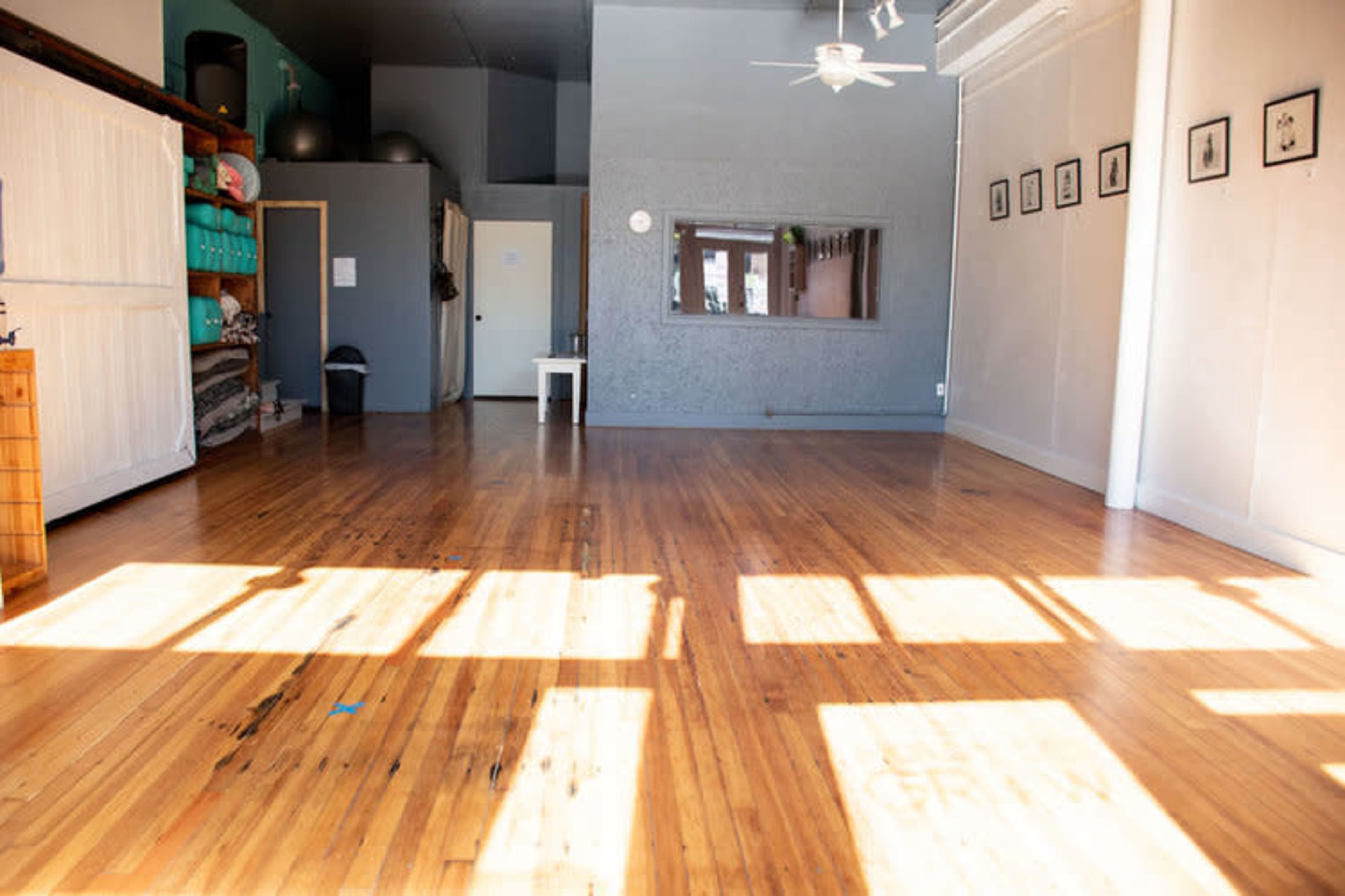 A spacious, sunlit room with wooden floors, a ceiling fan, and several framed pictures on the walls.