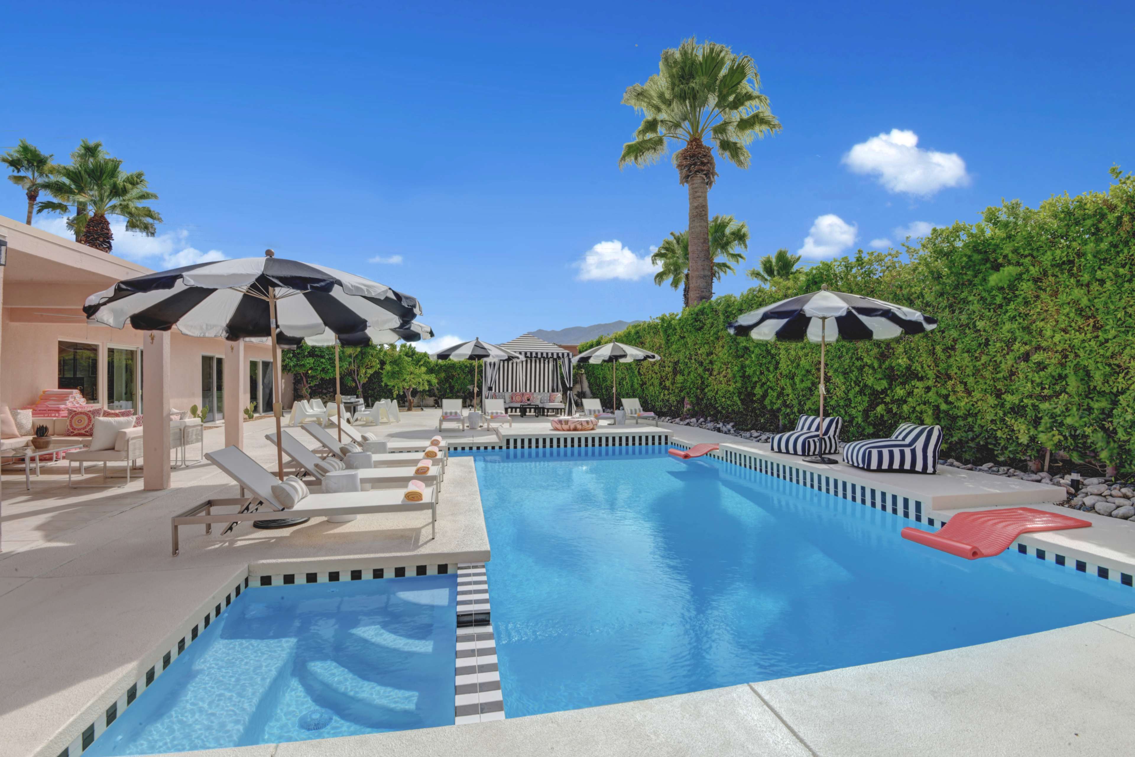 The image shows a swimming pool surrounded by lounge chairs and black-and-white striped umbrellas, with palm trees and hedges in the background.