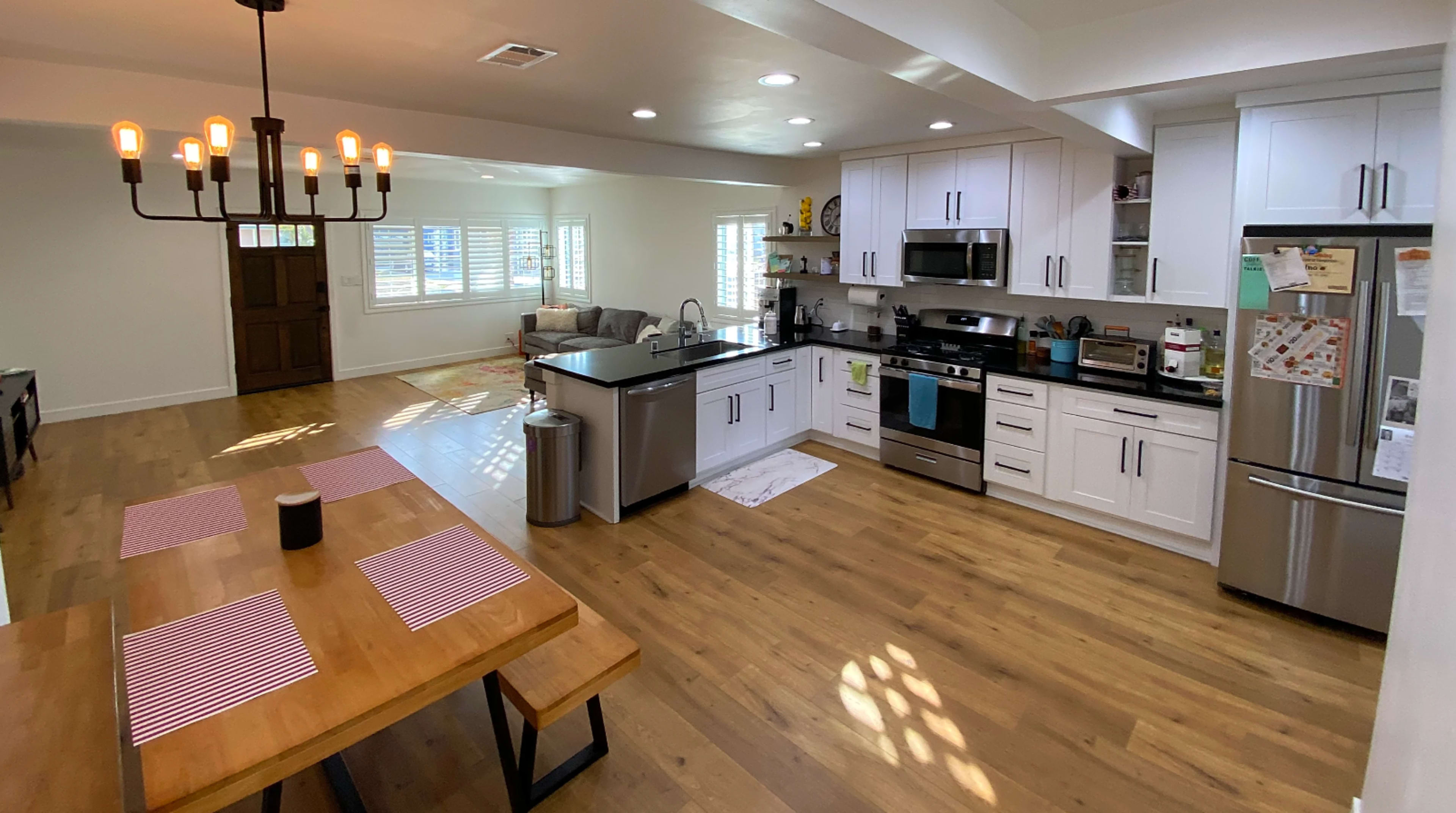 A modern kitchen and dining area with a large wooden table, white cabinetry, stainless steel appliances, and a living space in the background.