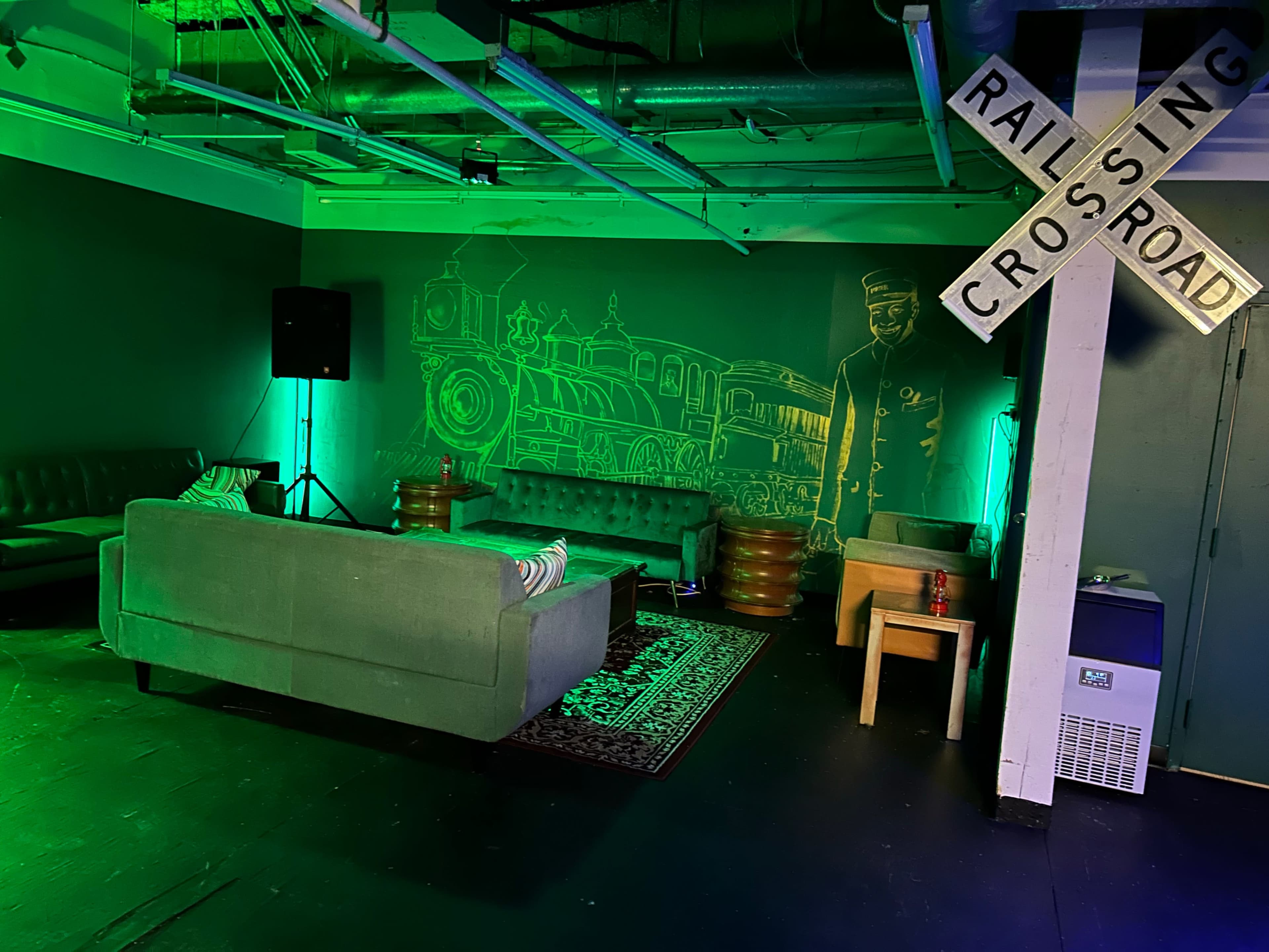 A lounge area with green couches, small wooden tables, and a mural of a train and conductor on the wall, alongside a railroad crossing sign.