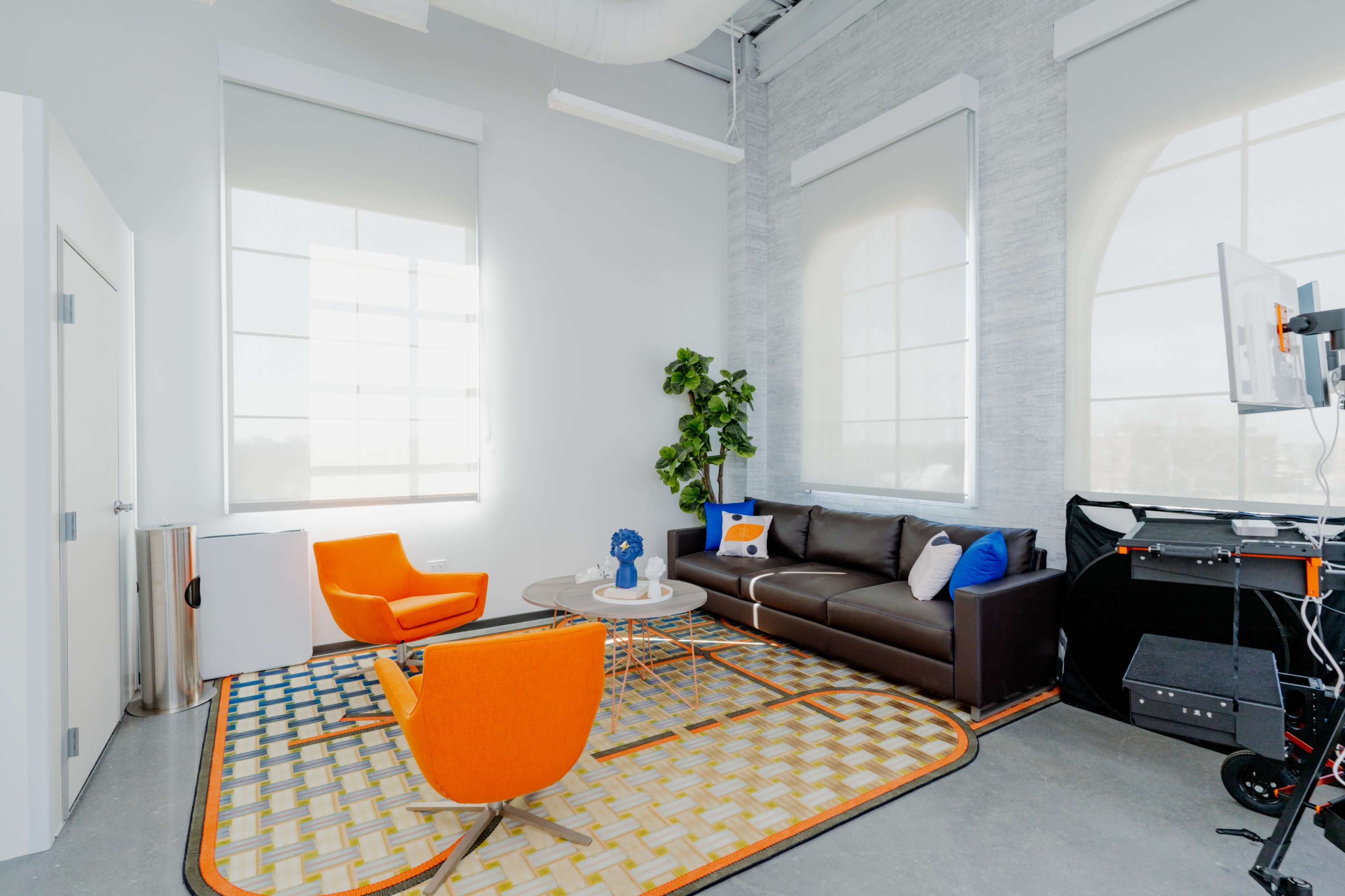 A modern office interior featuring a brown sofa, two orange chairs, a round coffee table, and a patterned area rug under bright, natural light from large windows.