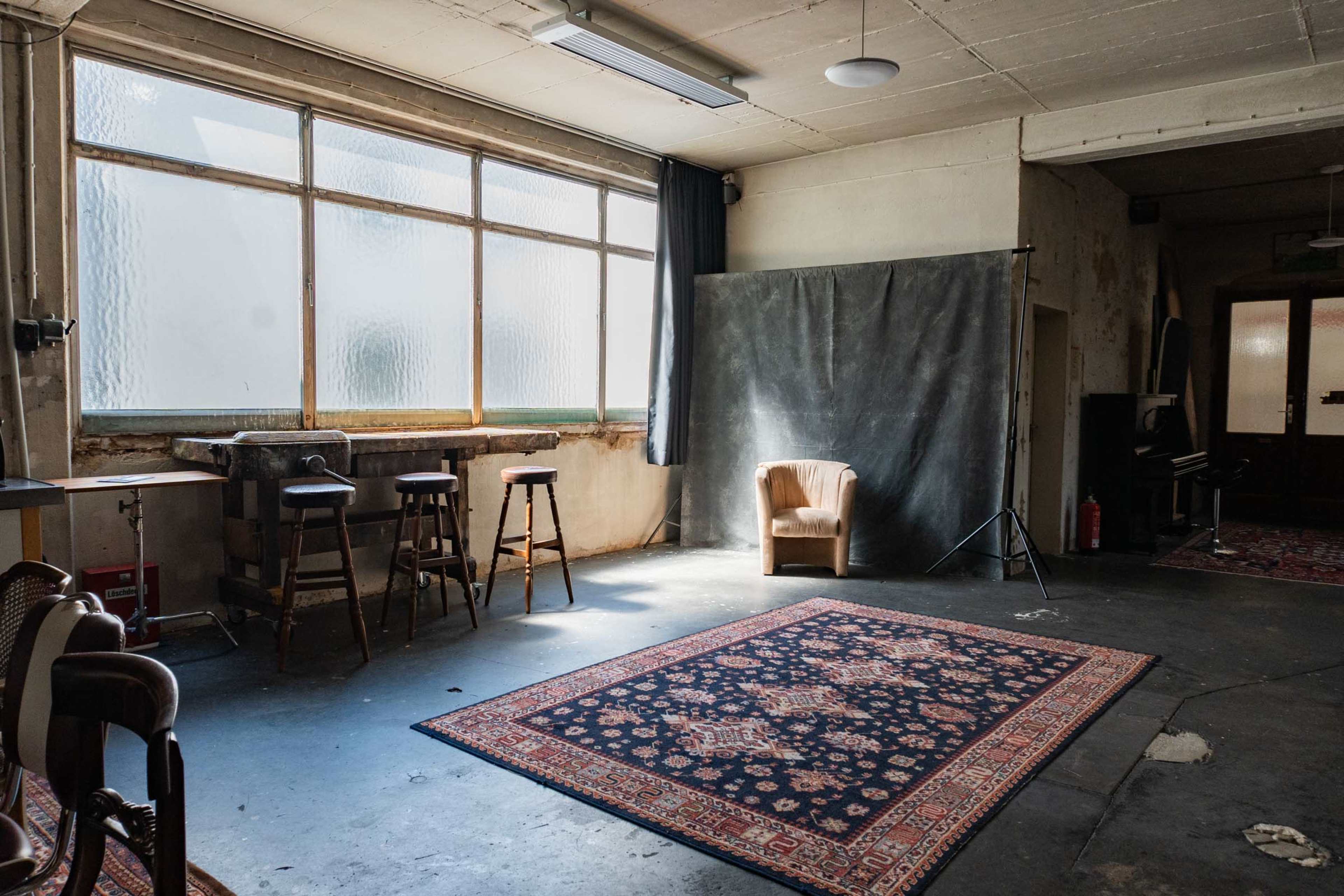 The image shows a spacious, industrial-style room with large windows, a vintage armchair, a rug on the floor, and a photography backdrop.
