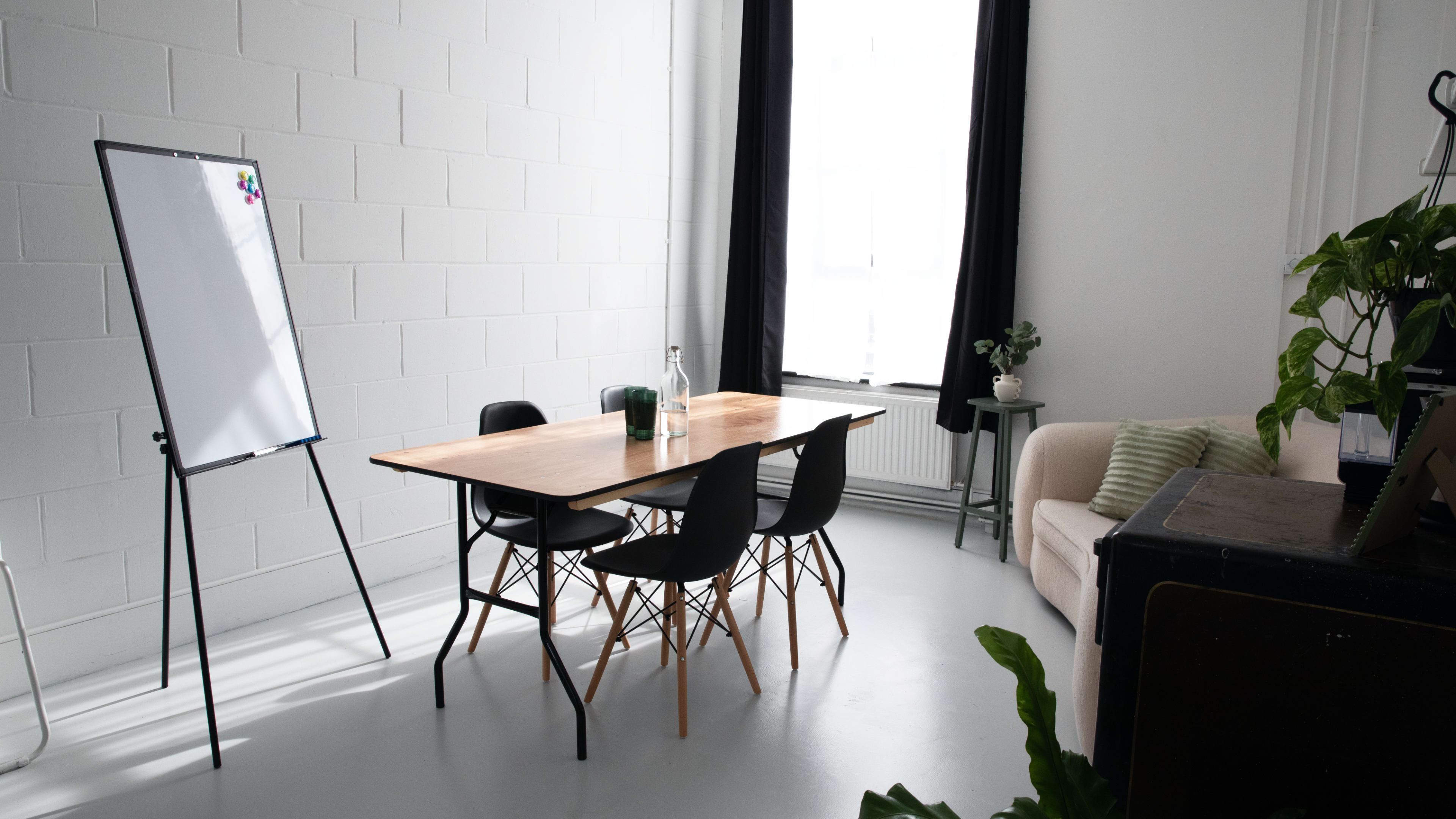 A bright, minimalist meeting room features a wooden table surrounded by black chairs, a whiteboard on an easel, and a sofa against the wall.