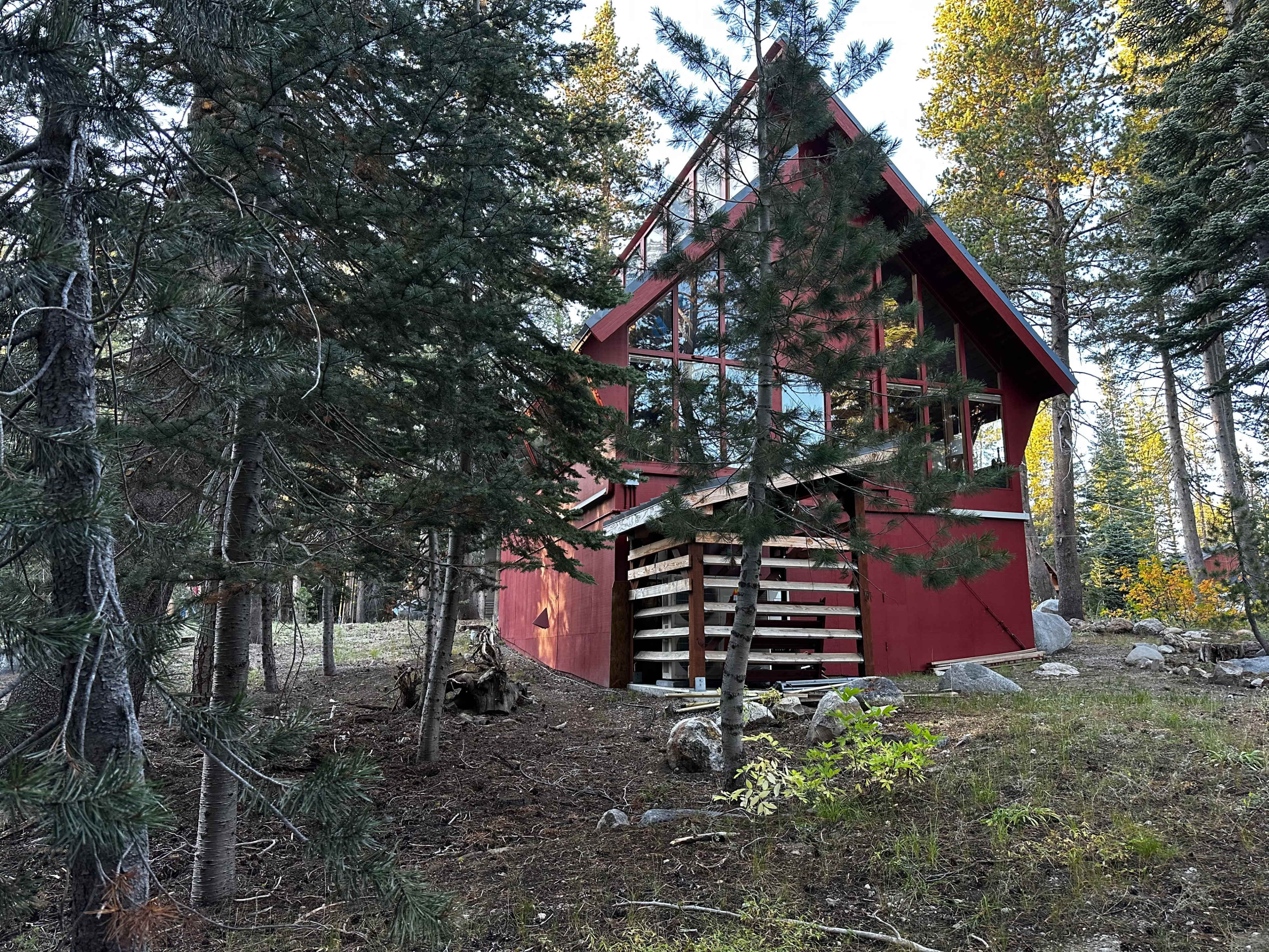 A red A-frame cabin with large windows is situated among tall trees and rocky terrain.