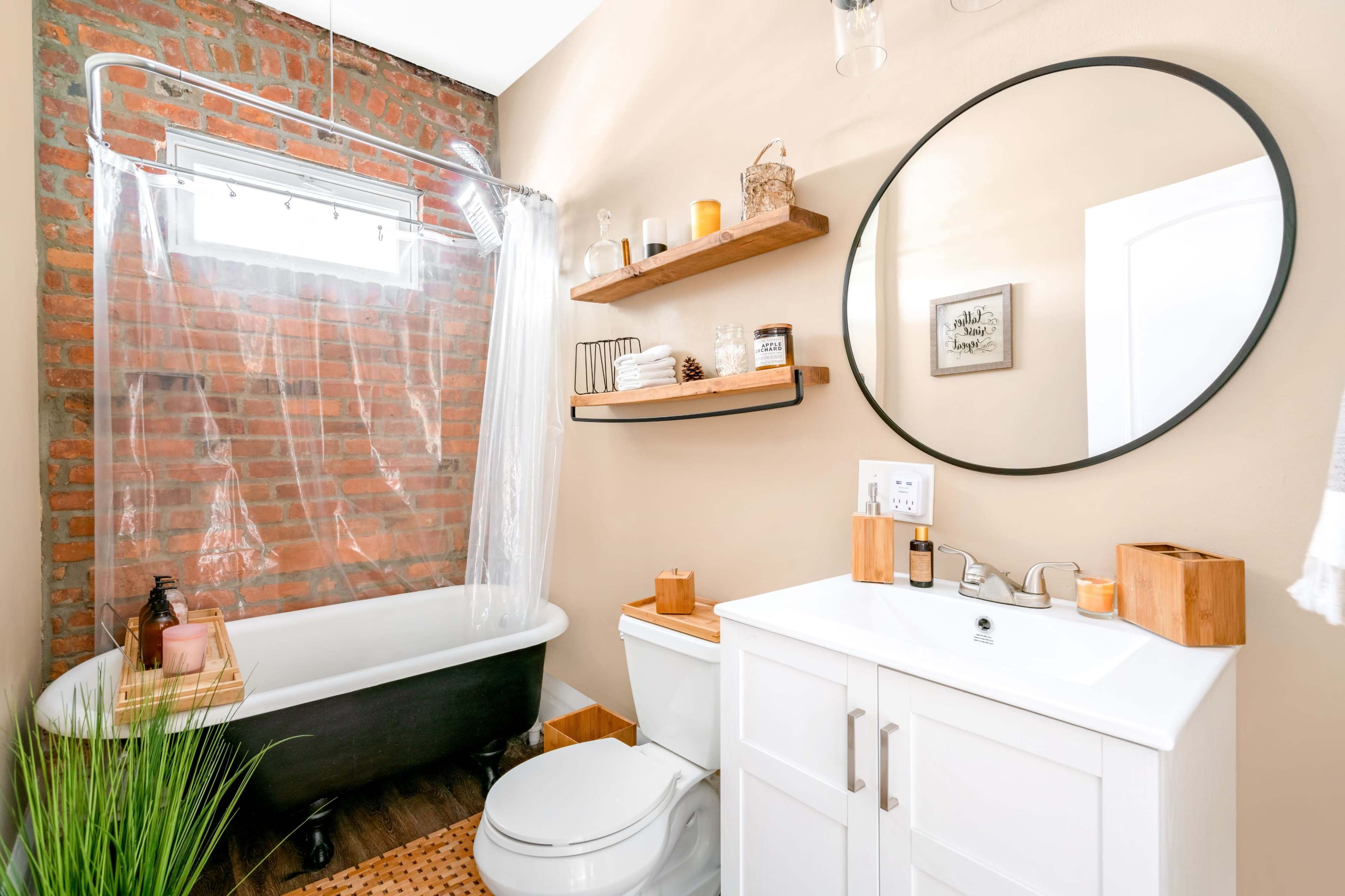 A bathroom featuring a black freestanding bathtub, a round mirror above a white vanity, and walls adorned with exposed brick and wooden shelves.