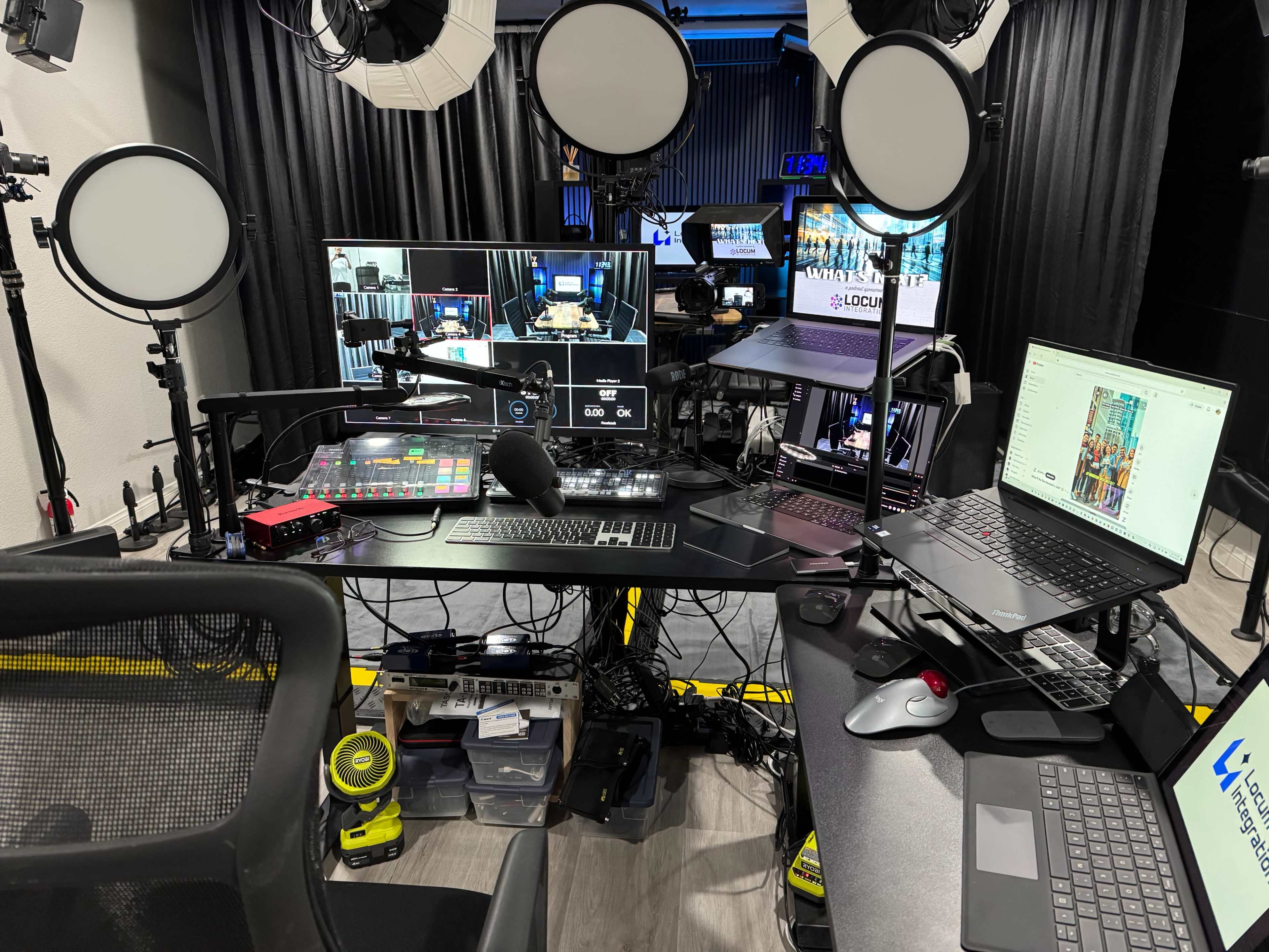 The image shows a multimedia production setup with multiple screens, professional lighting, and various tech equipment arranged on a desk.