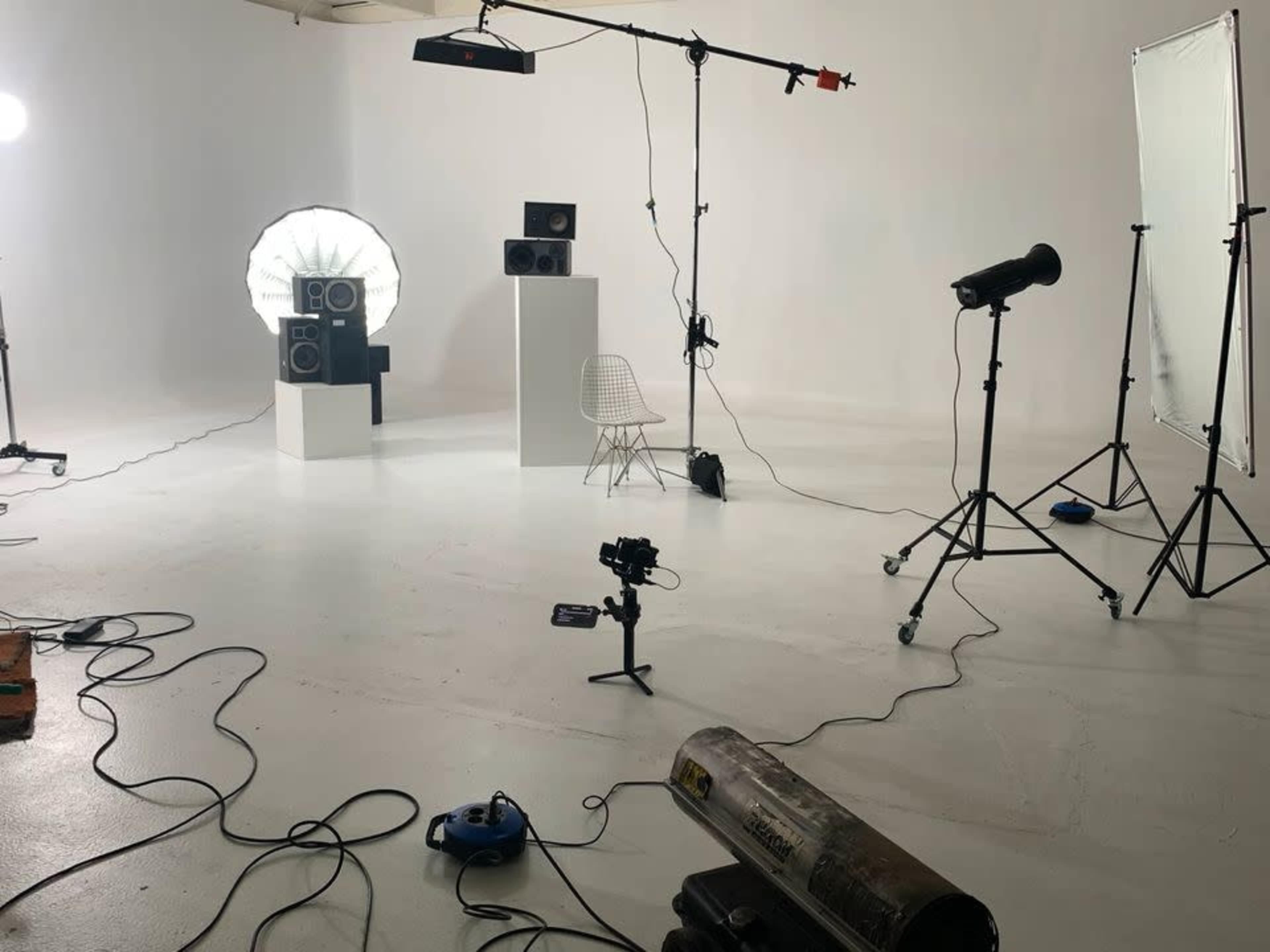 A photography studio with various lighting equipment, speakers on a pedestal, a white chair, and cables scattered on the floor.