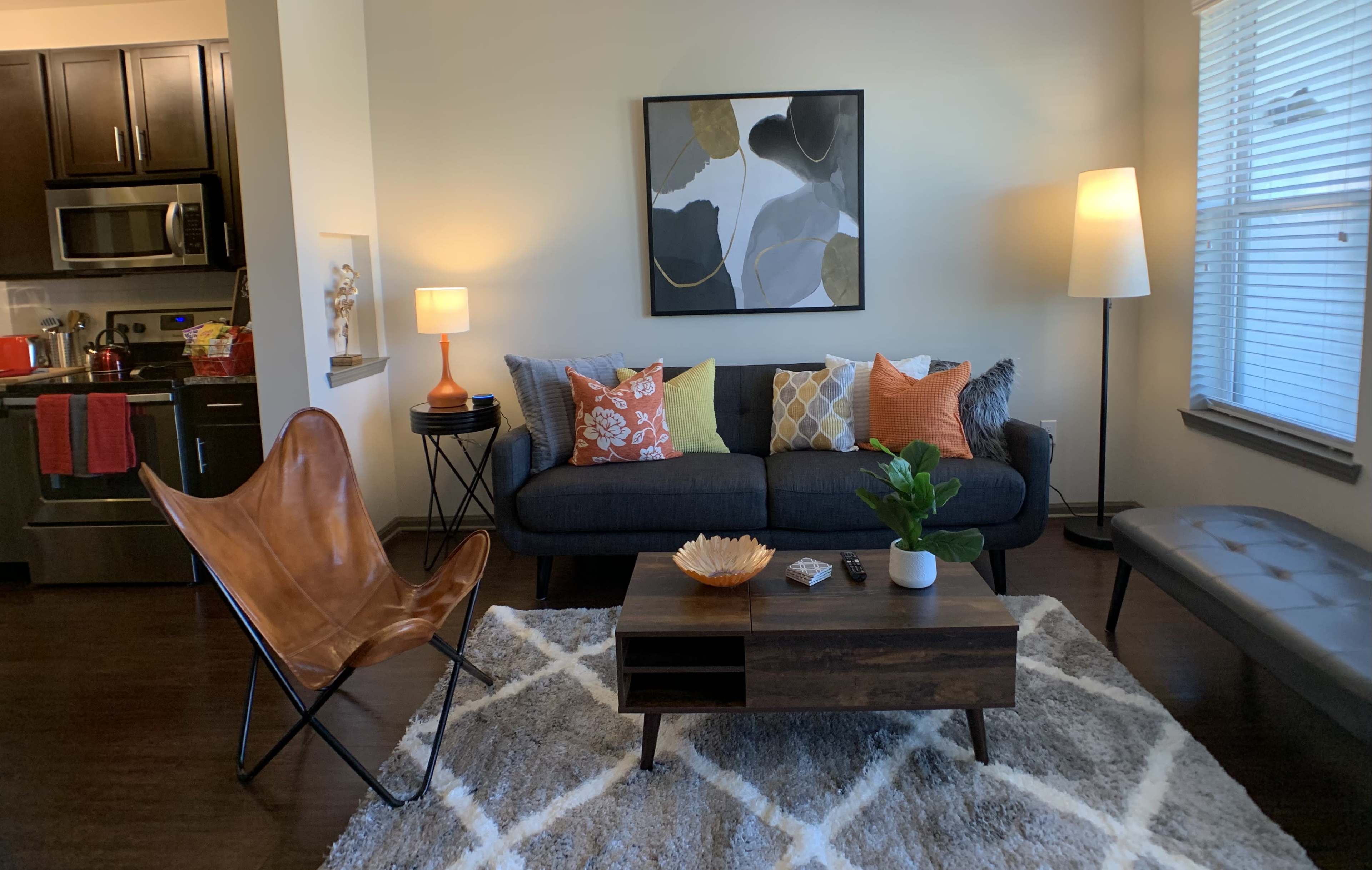 A living room with a dark sofa adorned with colorful pillows, a wooden coffee table, a decorative bowl, a plant, and a soft area rug, all set against light-colored walls and a window with blinds.