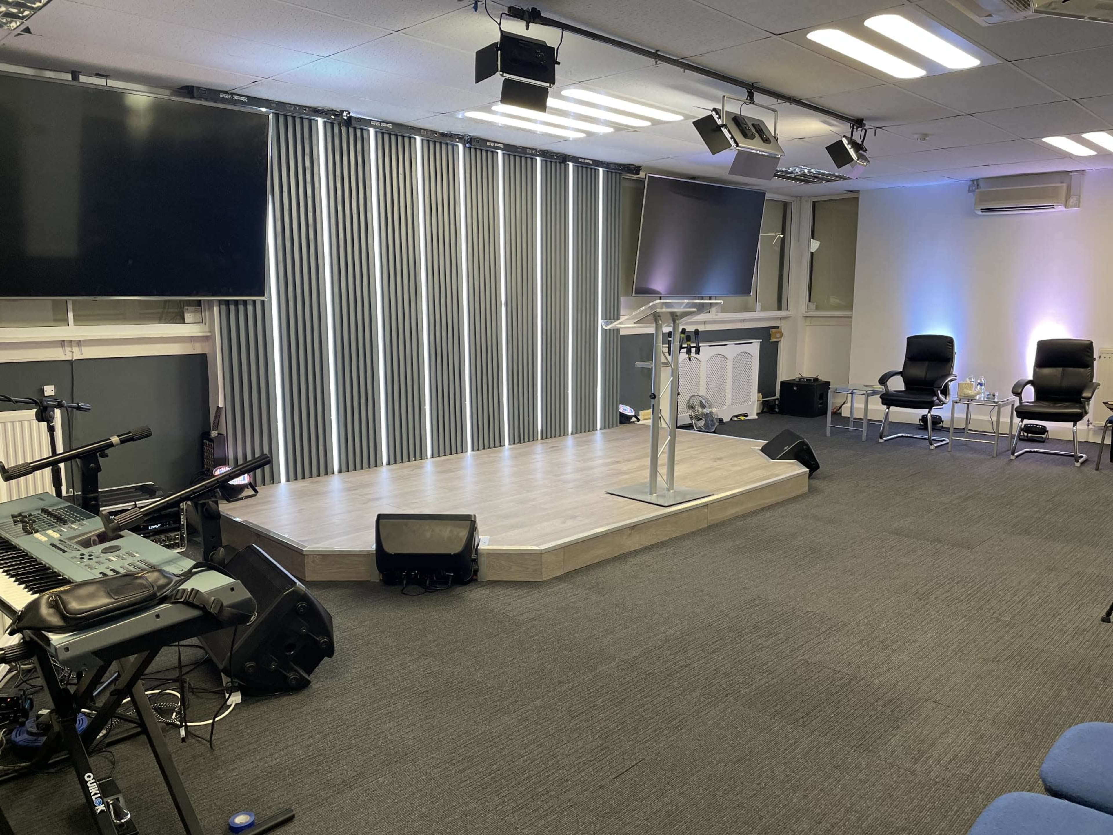 A stage setup in a room with two large screens, a podium, musical instruments, and seating arrangements.