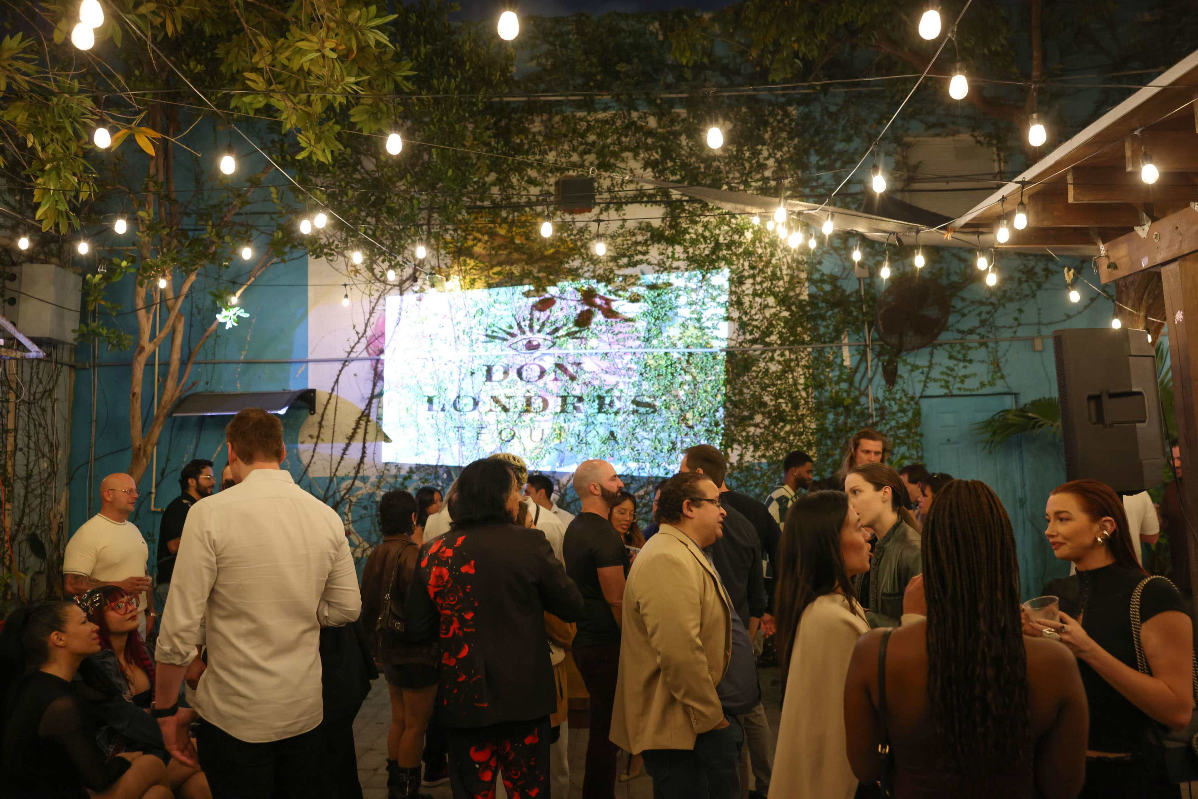 A large gathering of people socializes in an outdoor space adorned with string lights and greenery, featuring a projected display on a wall.