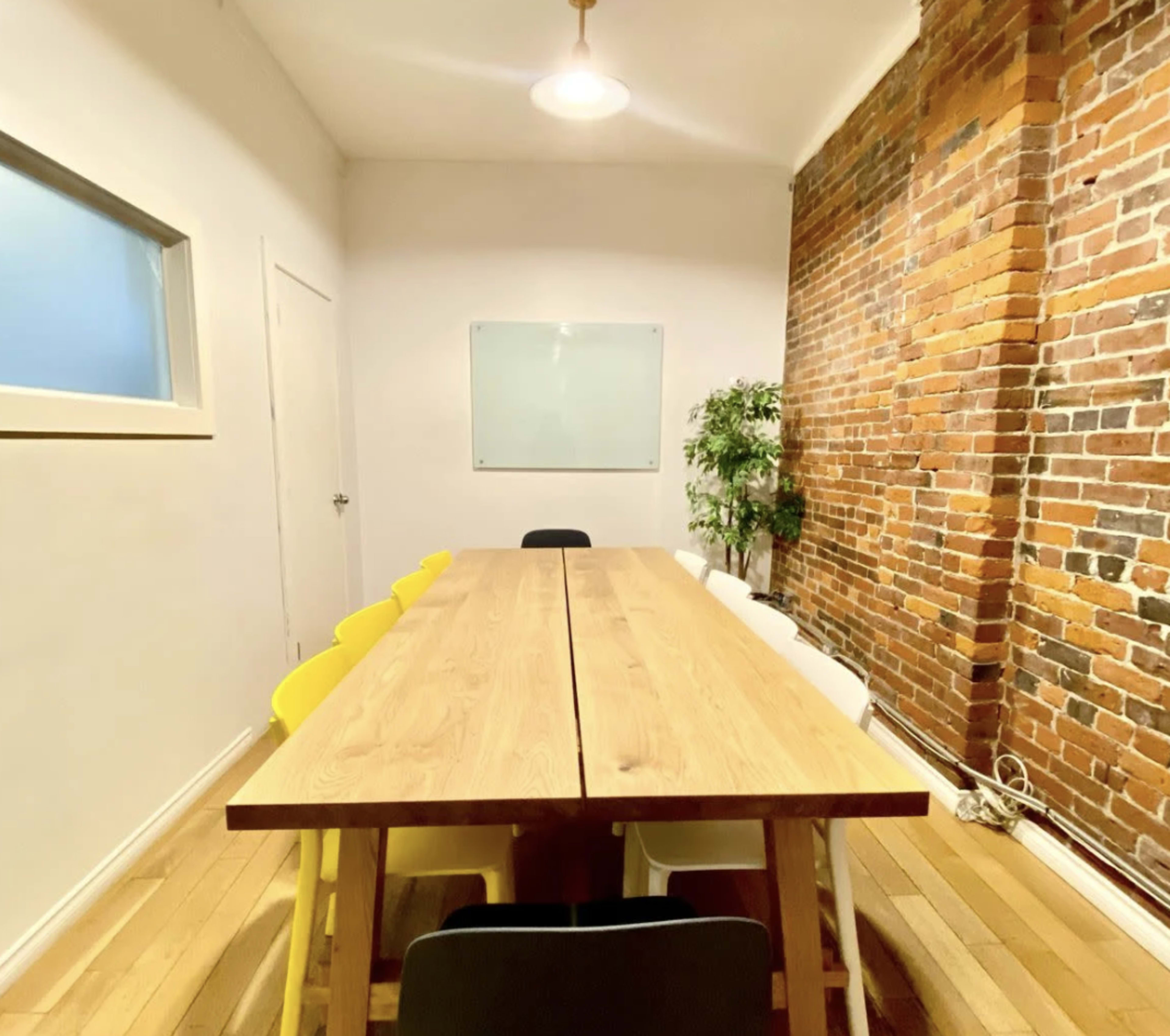 The image shows a conference room with a long wooden table surrounded by chairs of various colors, a brick wall on one side, and a whiteboard mounted on the wall.