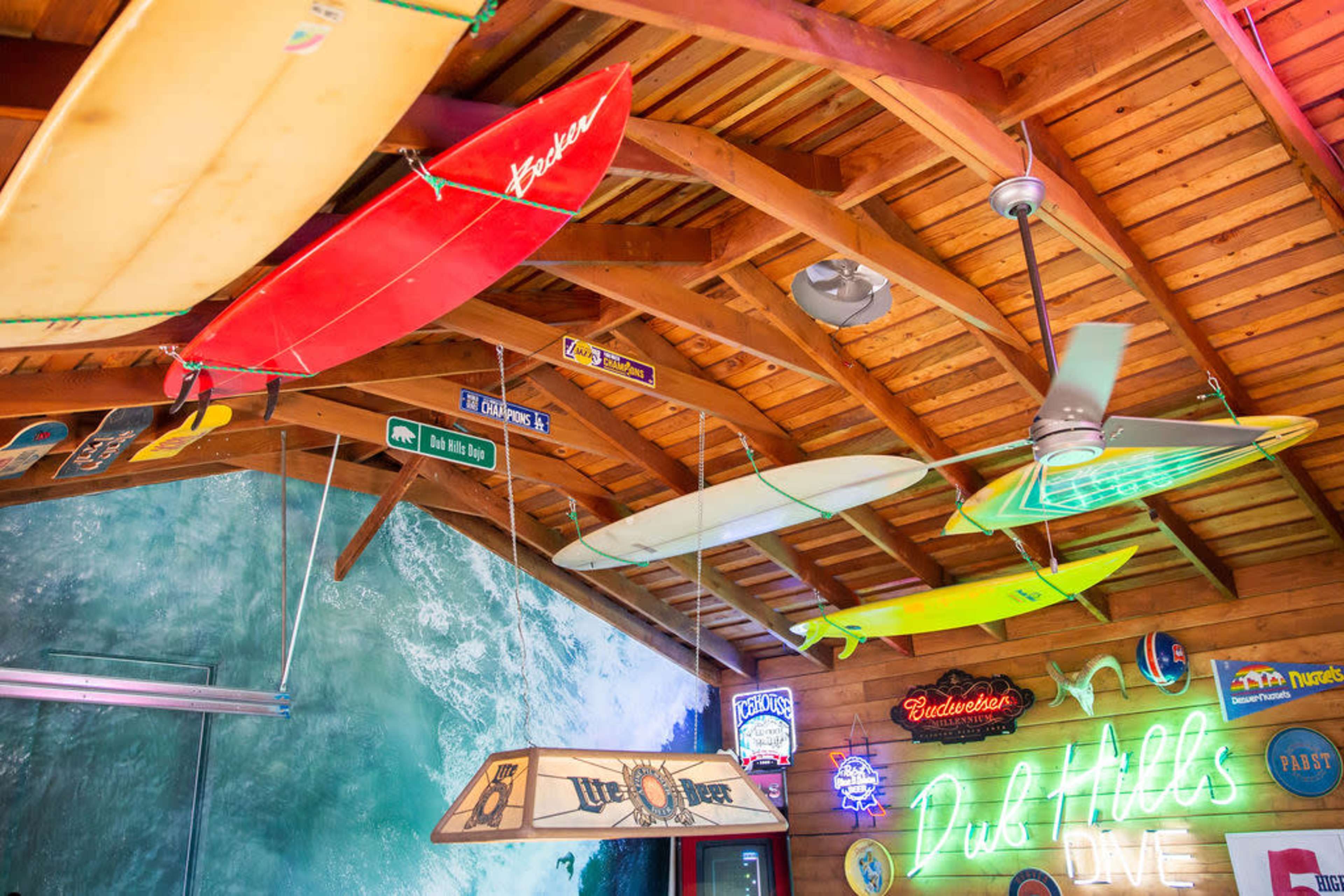 The interior of a bar features wooden beams adorned with colorful surfboards and neon signs.
