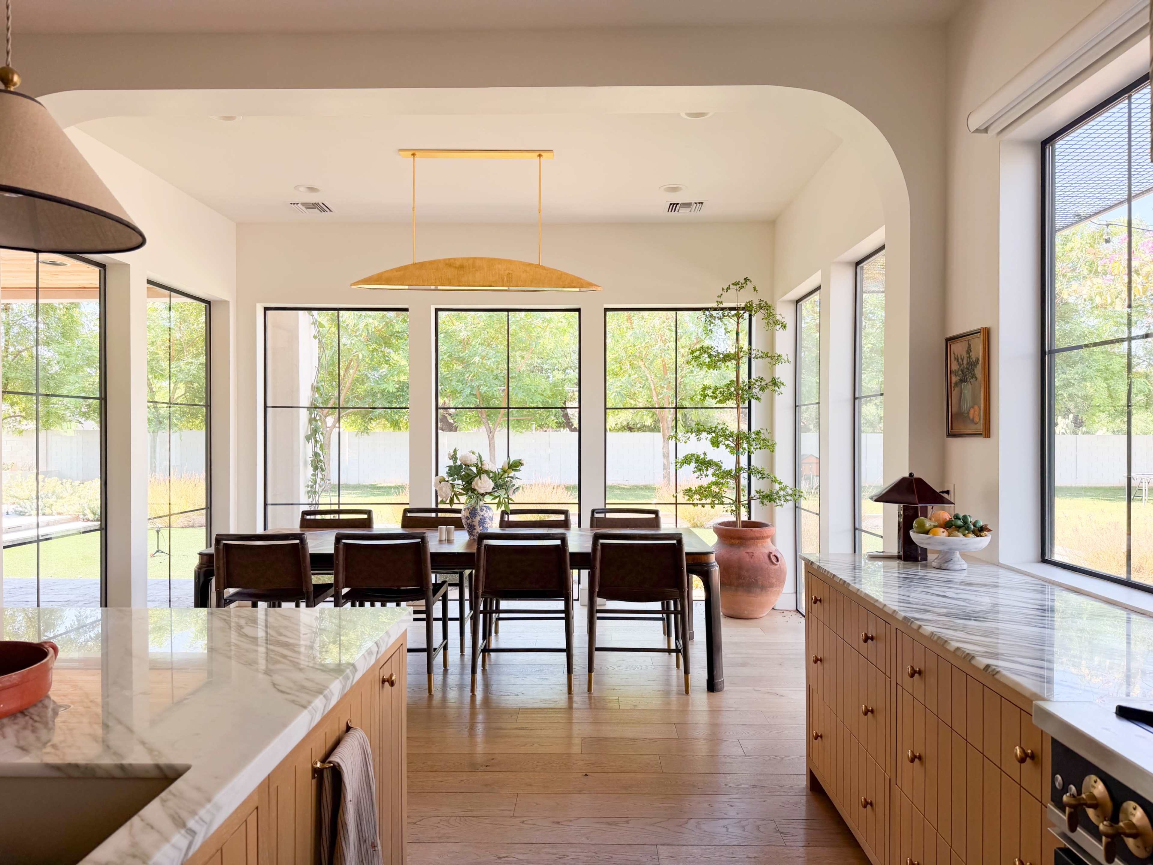 The image shows a modern dining area with a large table surrounded by chairs, illuminated by natural light coming through multiple large windows.
