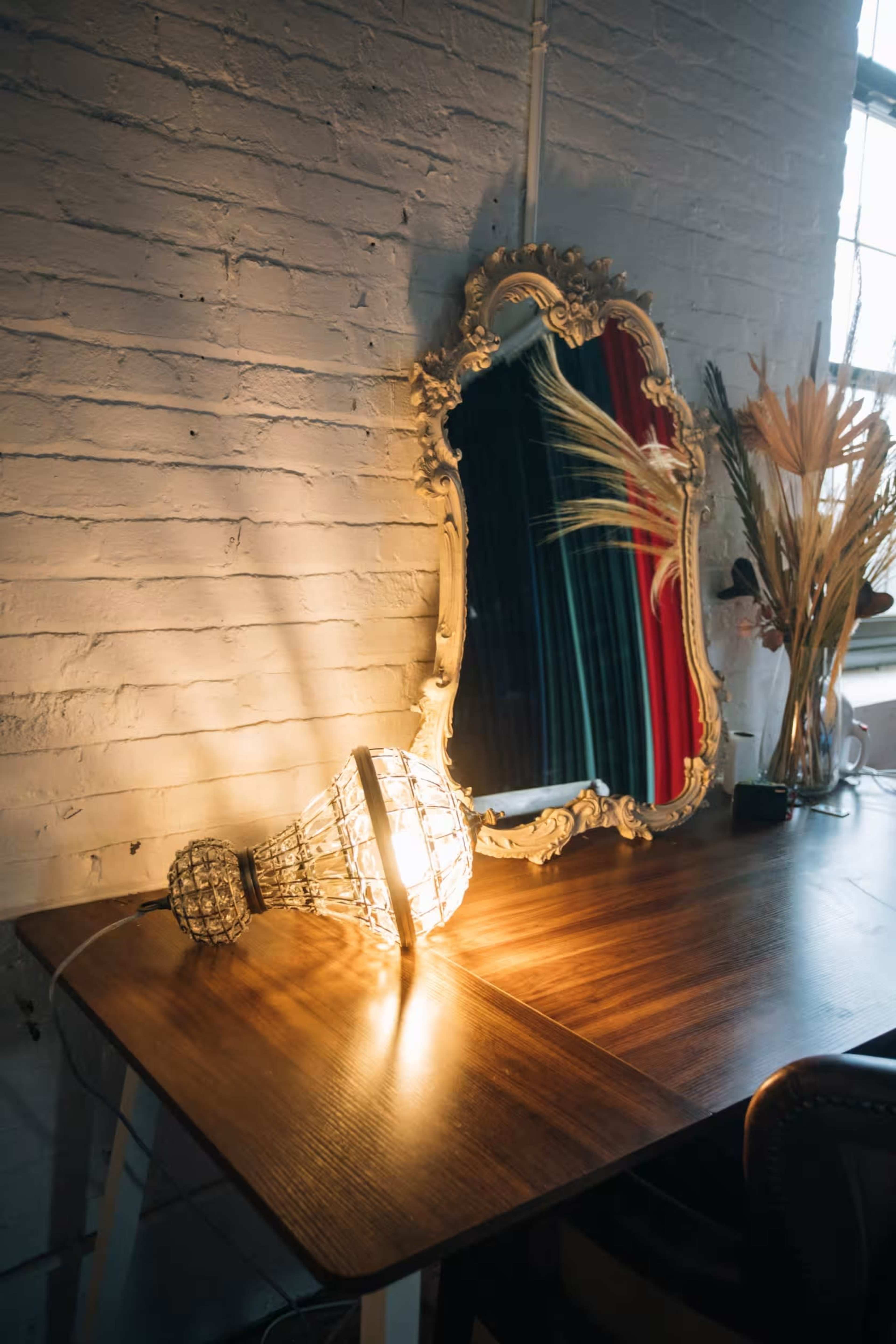A vintage mirror with a decorative frame reflects a light bulb lying on a wooden table beside dried plants.