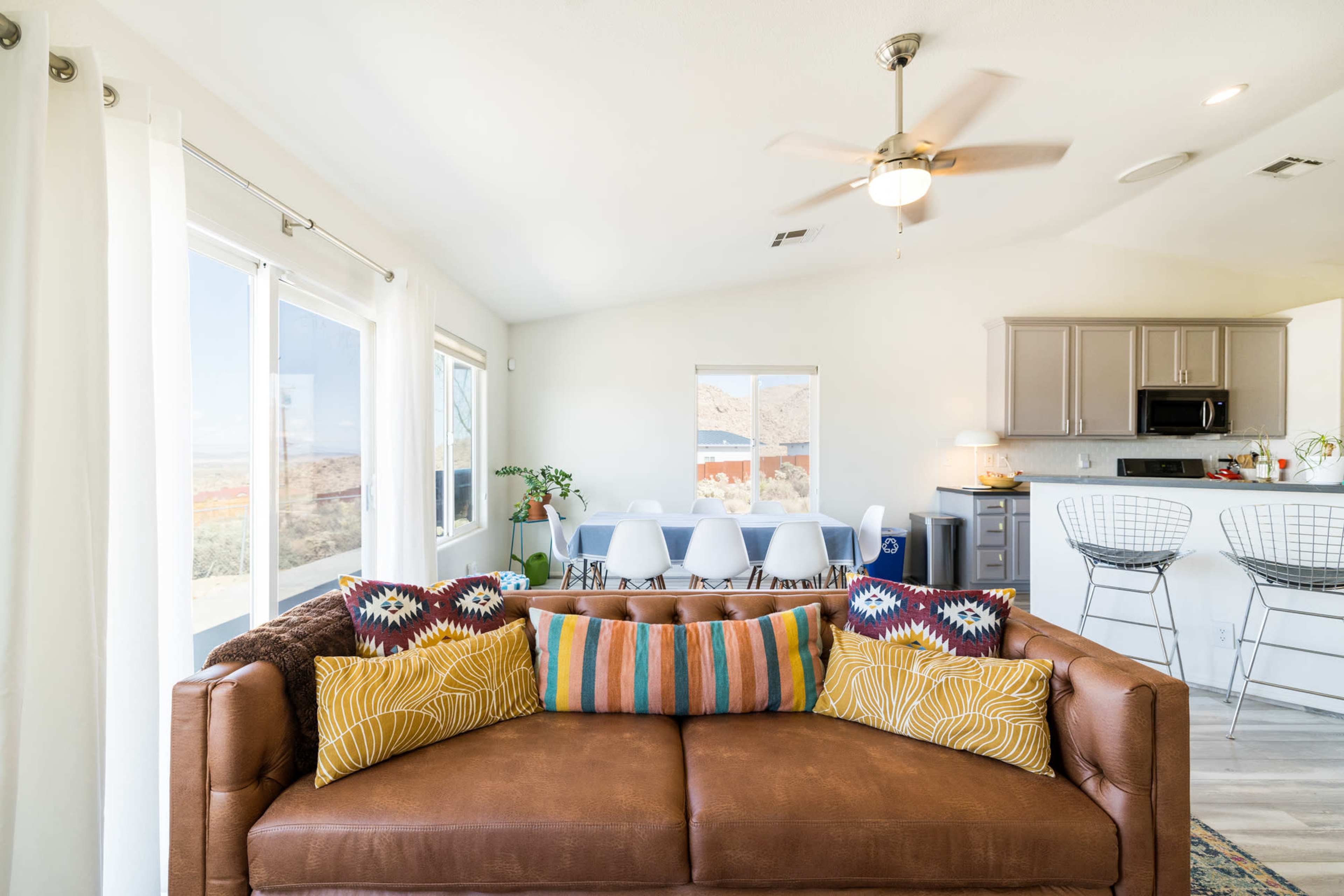 A living space features a brown sofa with colorful pillows in the foreground, and a dining area with a white table and chairs visible in the background.