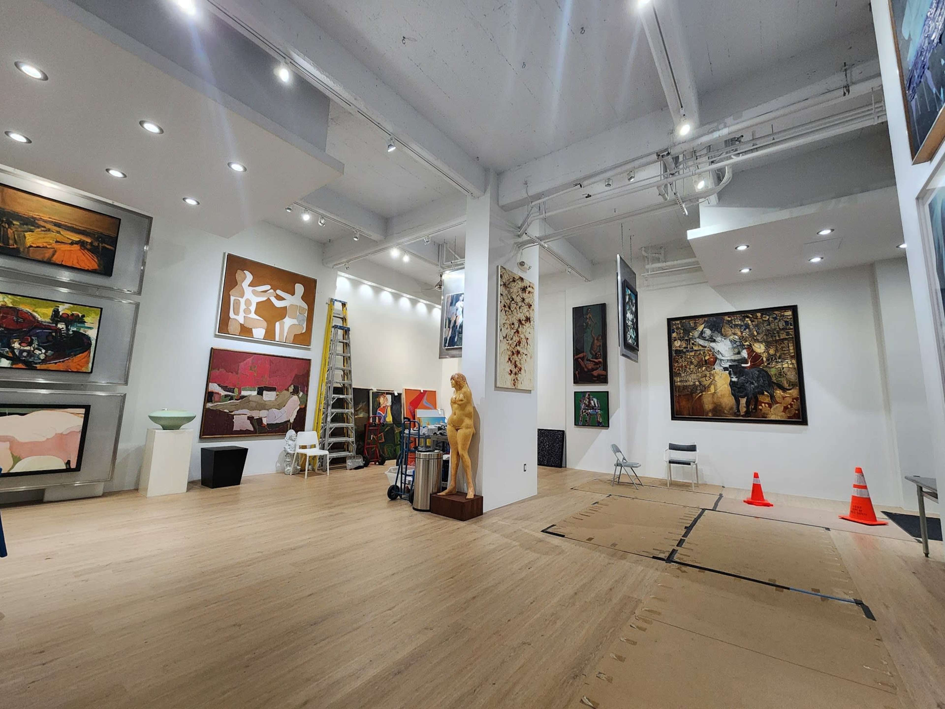 An art gallery with various paintings on the walls, wooden flooring, a statue, and construction barriers in the corner.